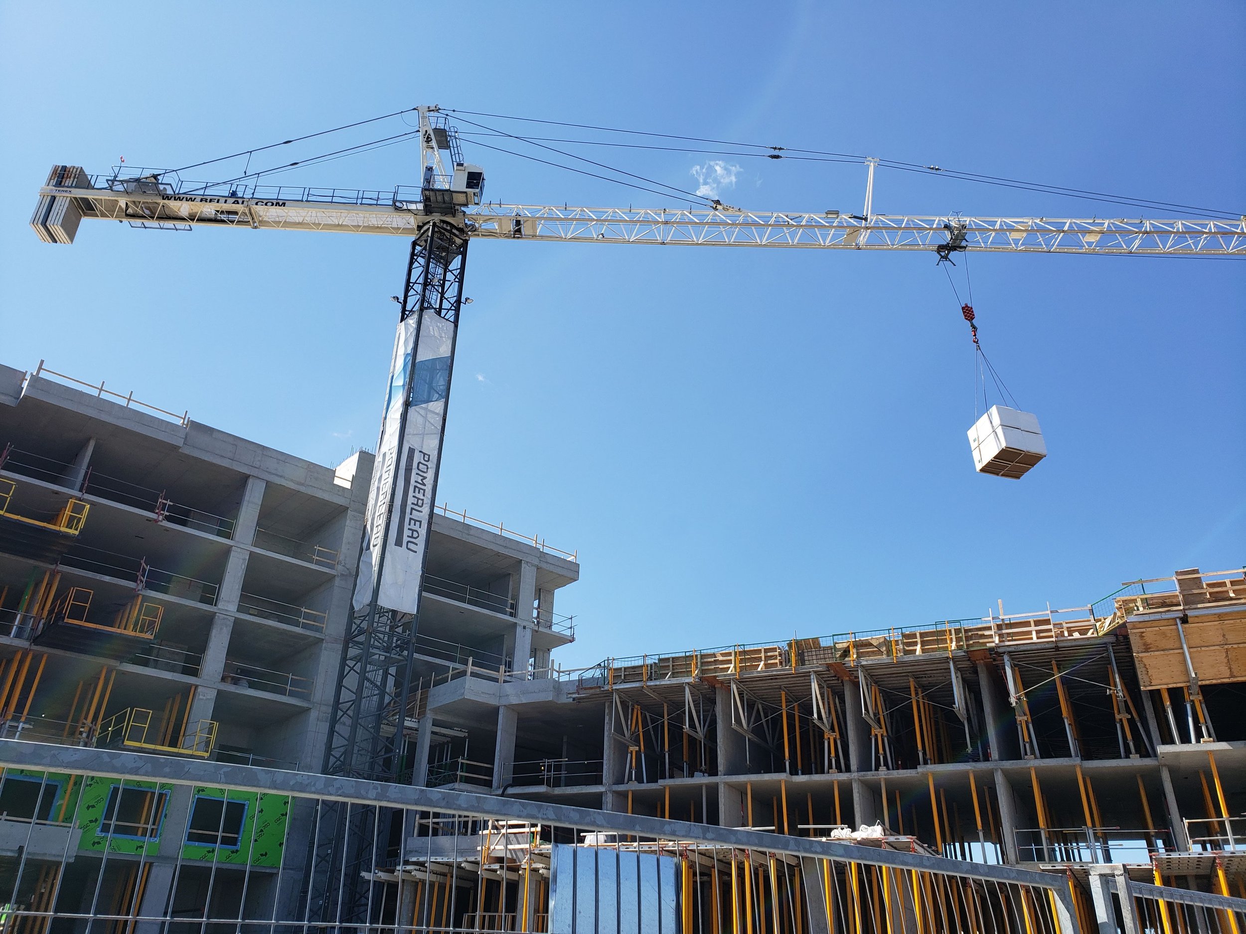 Construction site with a tall crane lifting materials, multiple floors of a building under construction, scaffolding, and building materials visible at the site, with a clear blue sky in the background.
