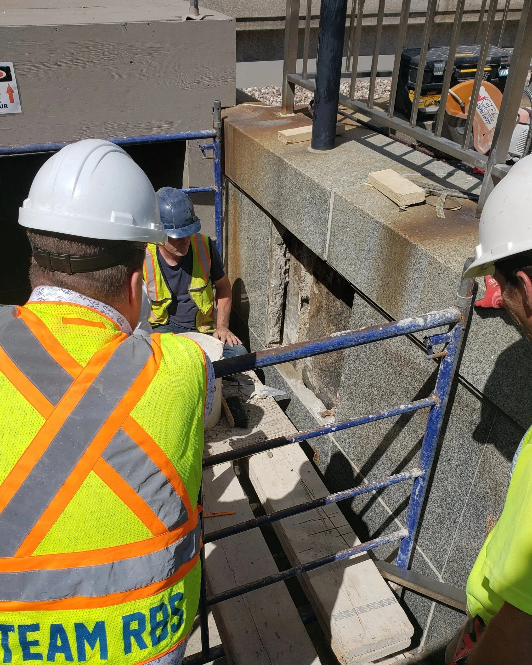 Construction workers wearing safety helmets and vests working at a building site, with scaffolding and construction tools visible.