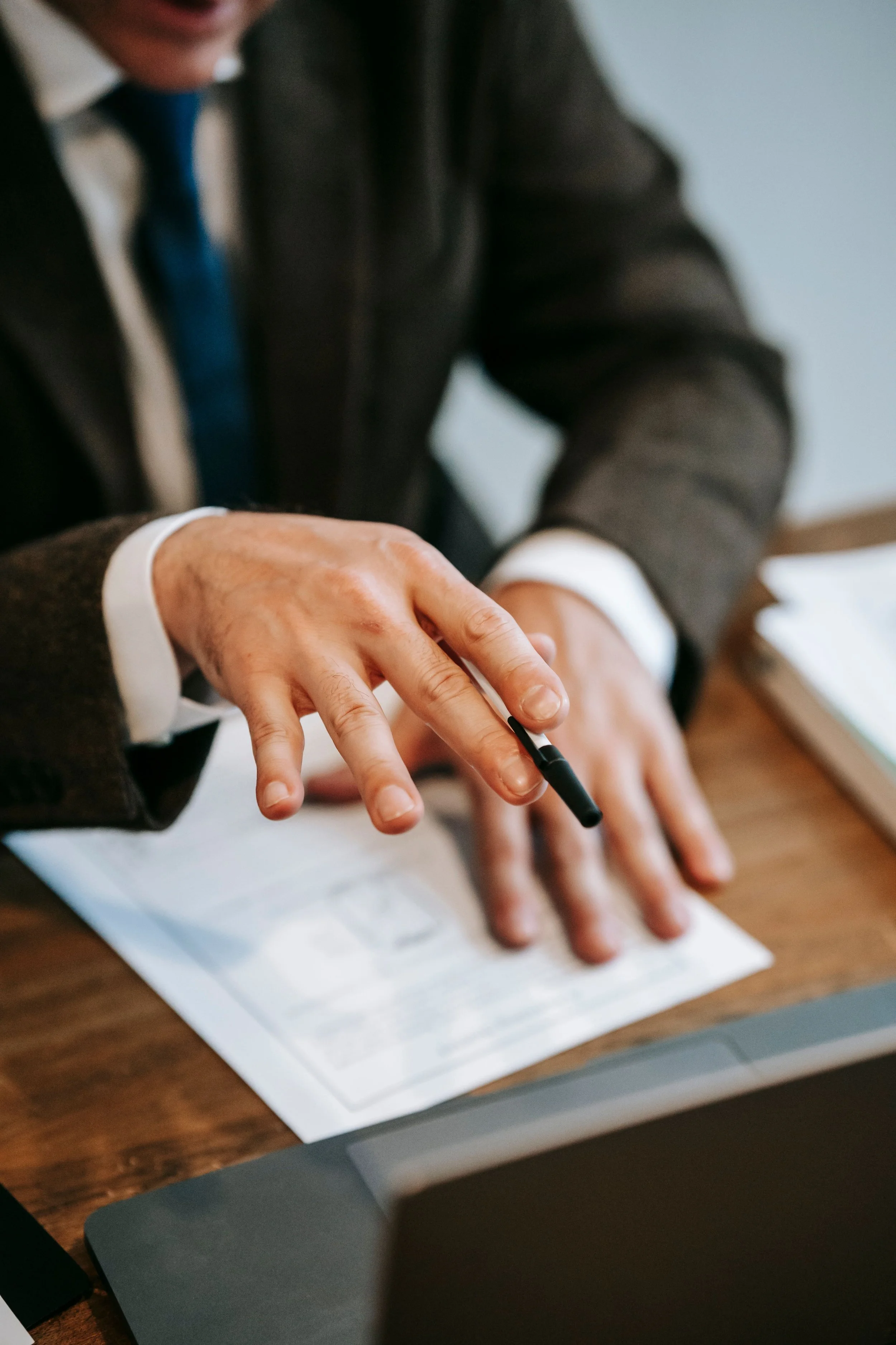 Close-up of a person's hand and fingers, holding a pen above a piece of paper on a wooden desk. The person is wearing a dark suit and a blue tie. The background is slightly blurred, showing part of the person's face and a light blue wall.