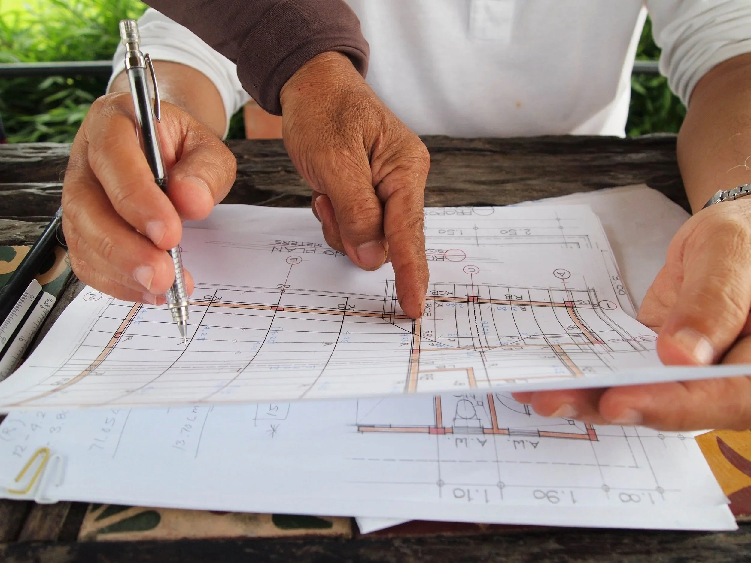 A person is drawing on blueprints or architectural plans with a pen, pointing at specific areas on the plans. The plans include technical drawings and measurements.
