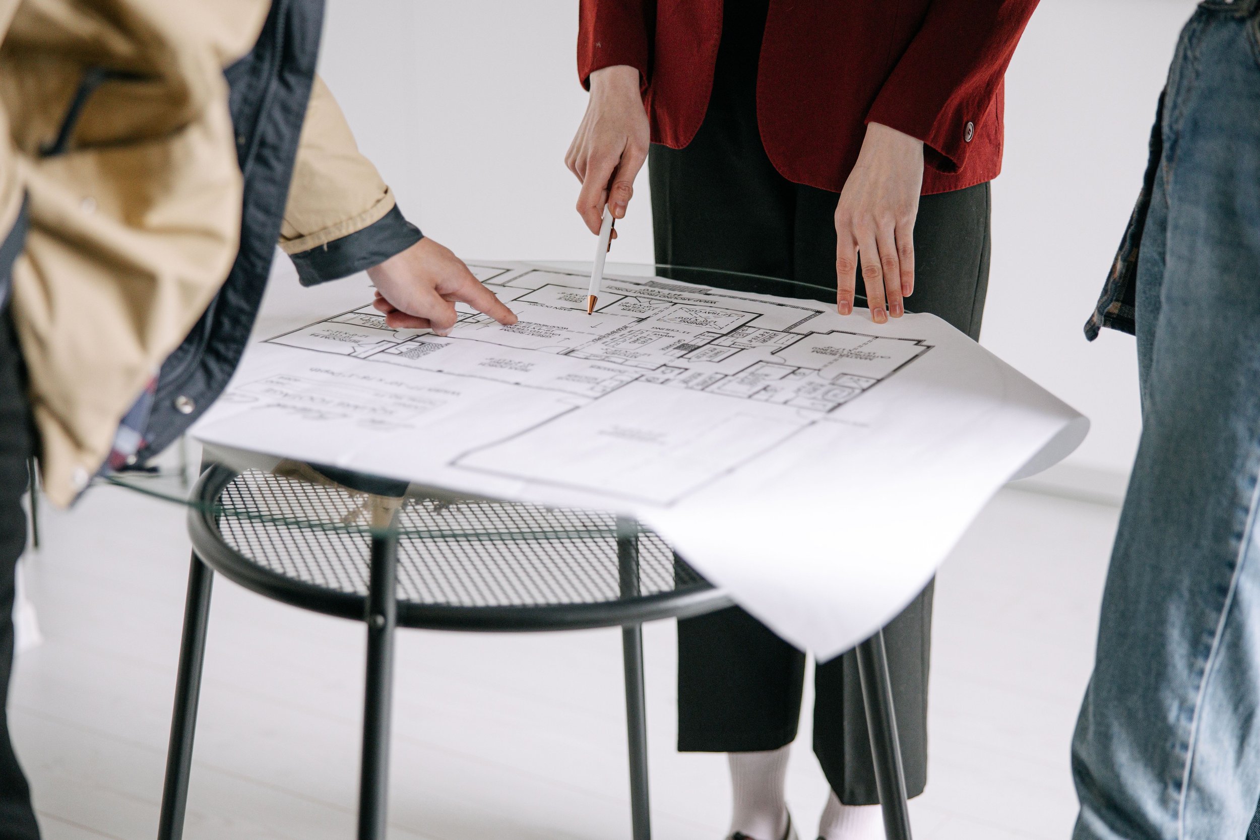 People gathered around a table examining architectural blueprints, with their hands pointing and holding pens, in a modern office setting.