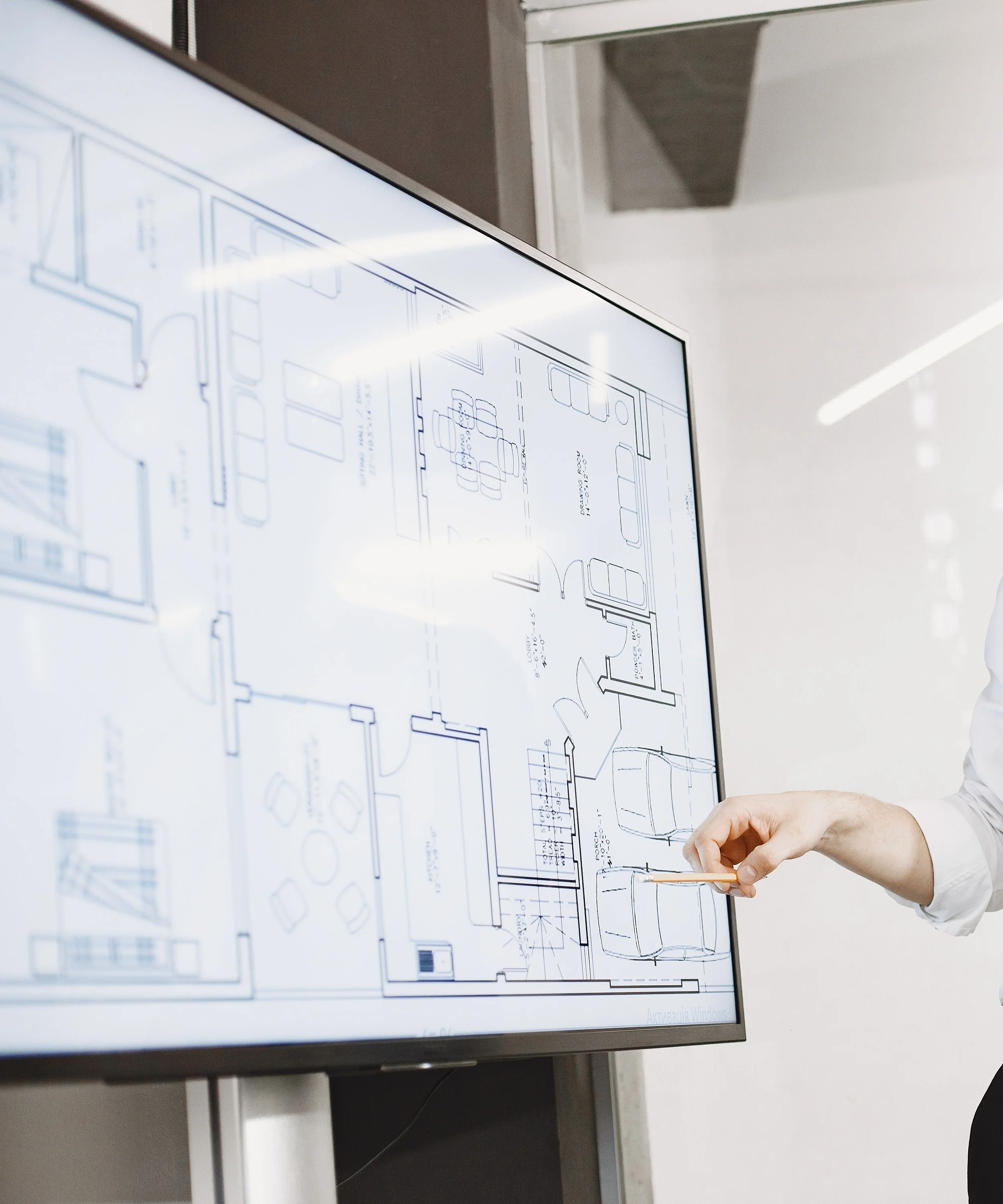 A person is standing next to a large display screen showing architectural blueprints and floor plans, using a pencil or pointing device.