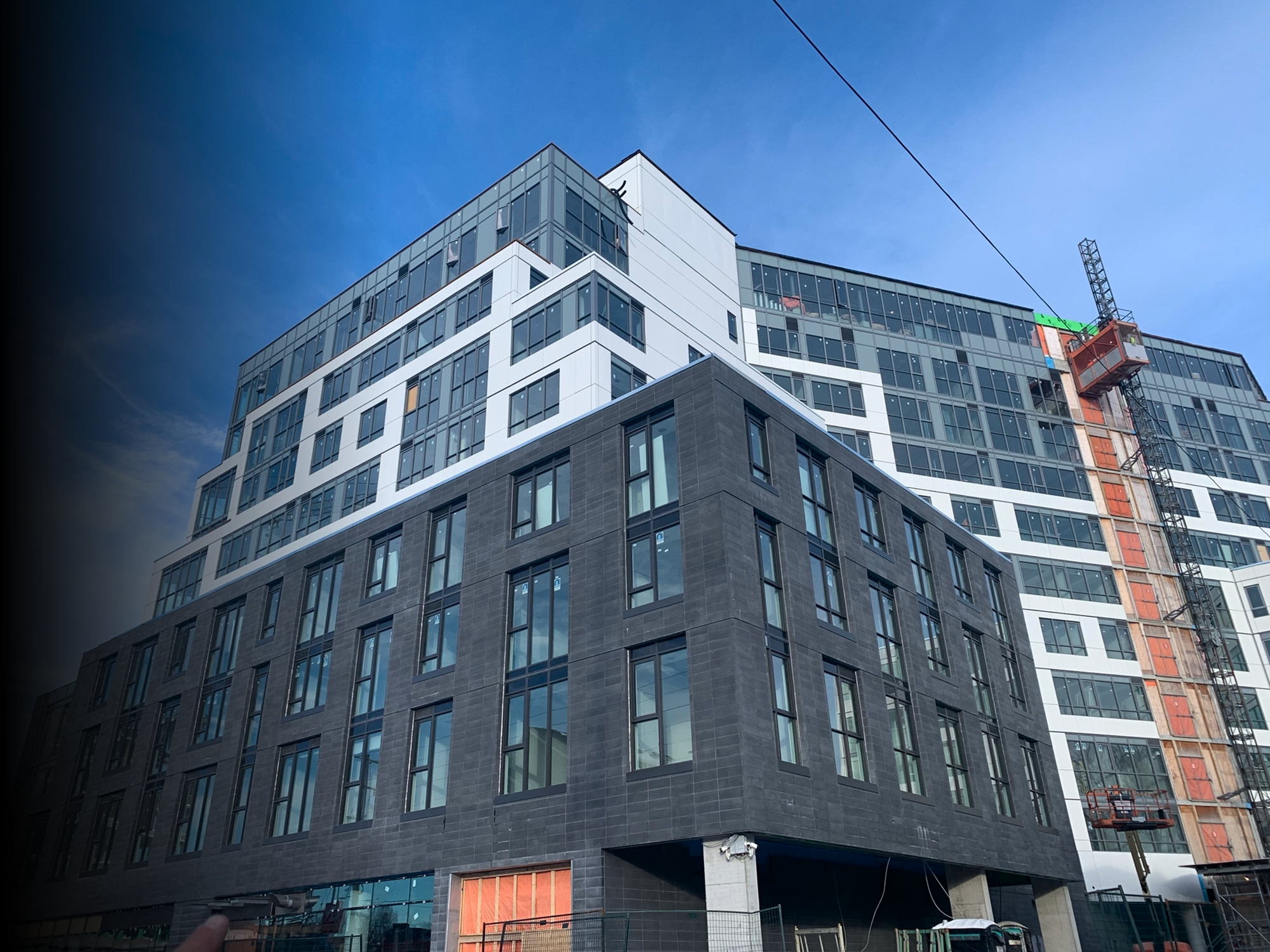 A modern multi-story building under construction with glass windows and black and white exterior panels. A construction crane is visible on the right side, and parts of the building are still under construction with exposed brickwork and scaffolding.