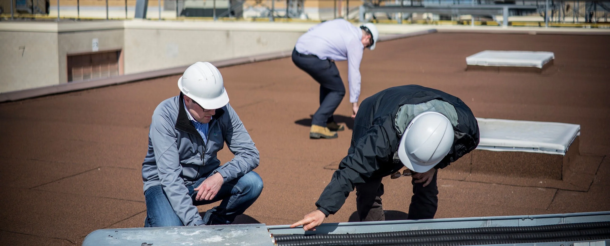 Three construction workers wearing safety helmets inspecting a rooftop from different positions, with one squatting, one leaning forward, and another bending down.