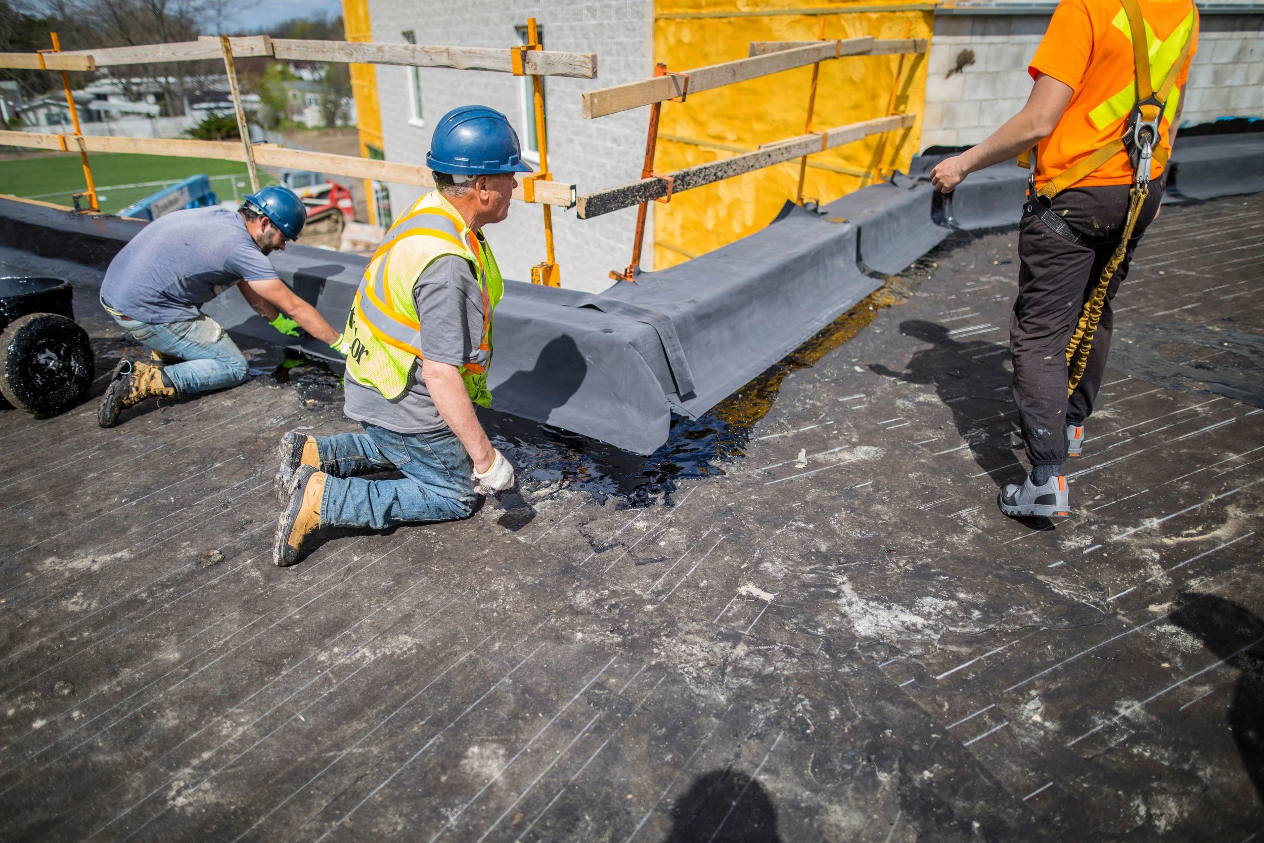 Construction workers installing roofing materials on a house. Two workers are kneeling and standing on the roof, with one applying roofing underlayment and another preparing the materials. They are wearing helmets and high-visibility safety vests.