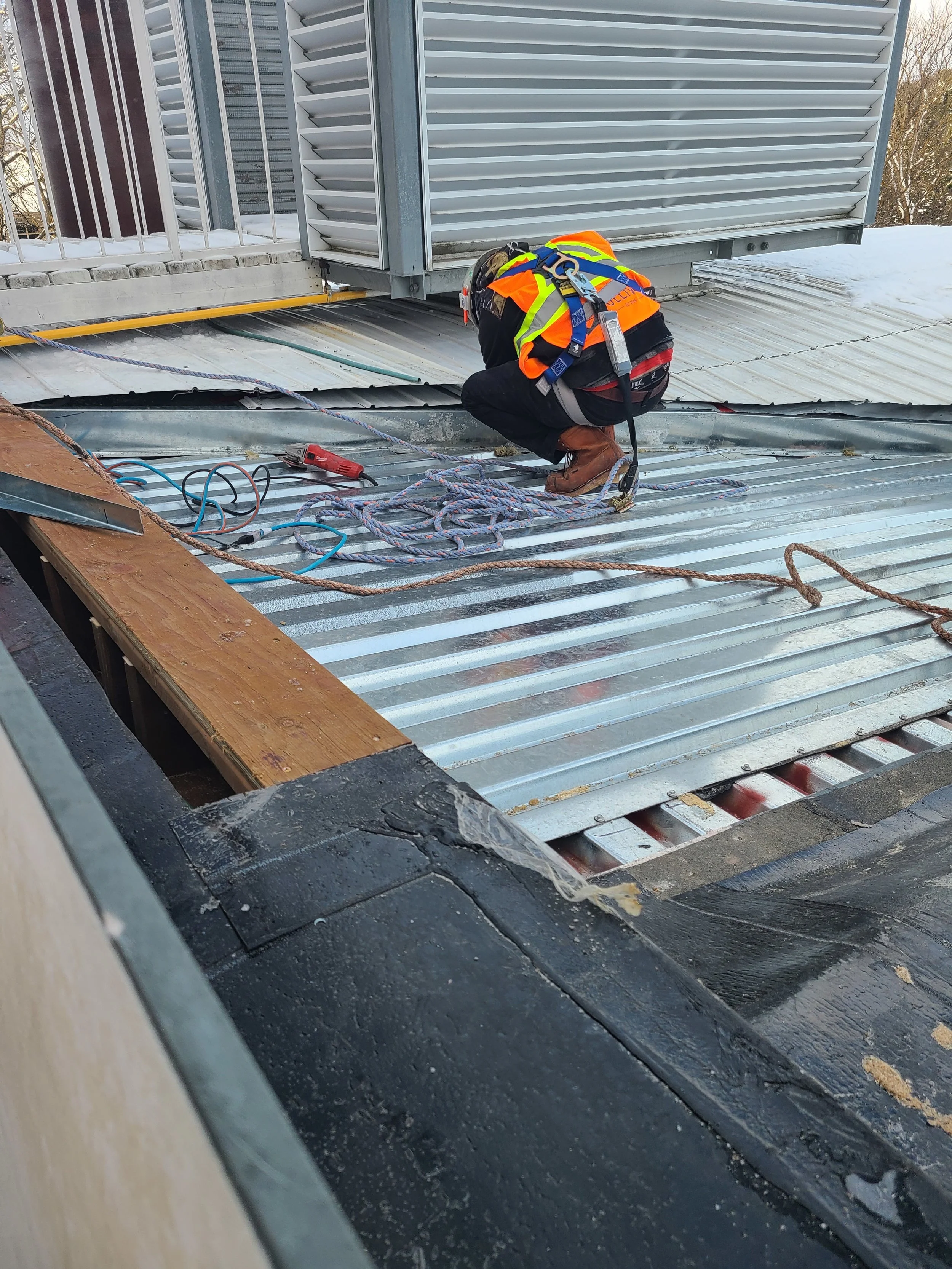 A construction worker wearing a safety vest, helmet, and harness is kneeling on a metal rooftop, working on a vent or HVAC unit. There are ropes, tools, and wooden planks around him, with snow visible in the background.