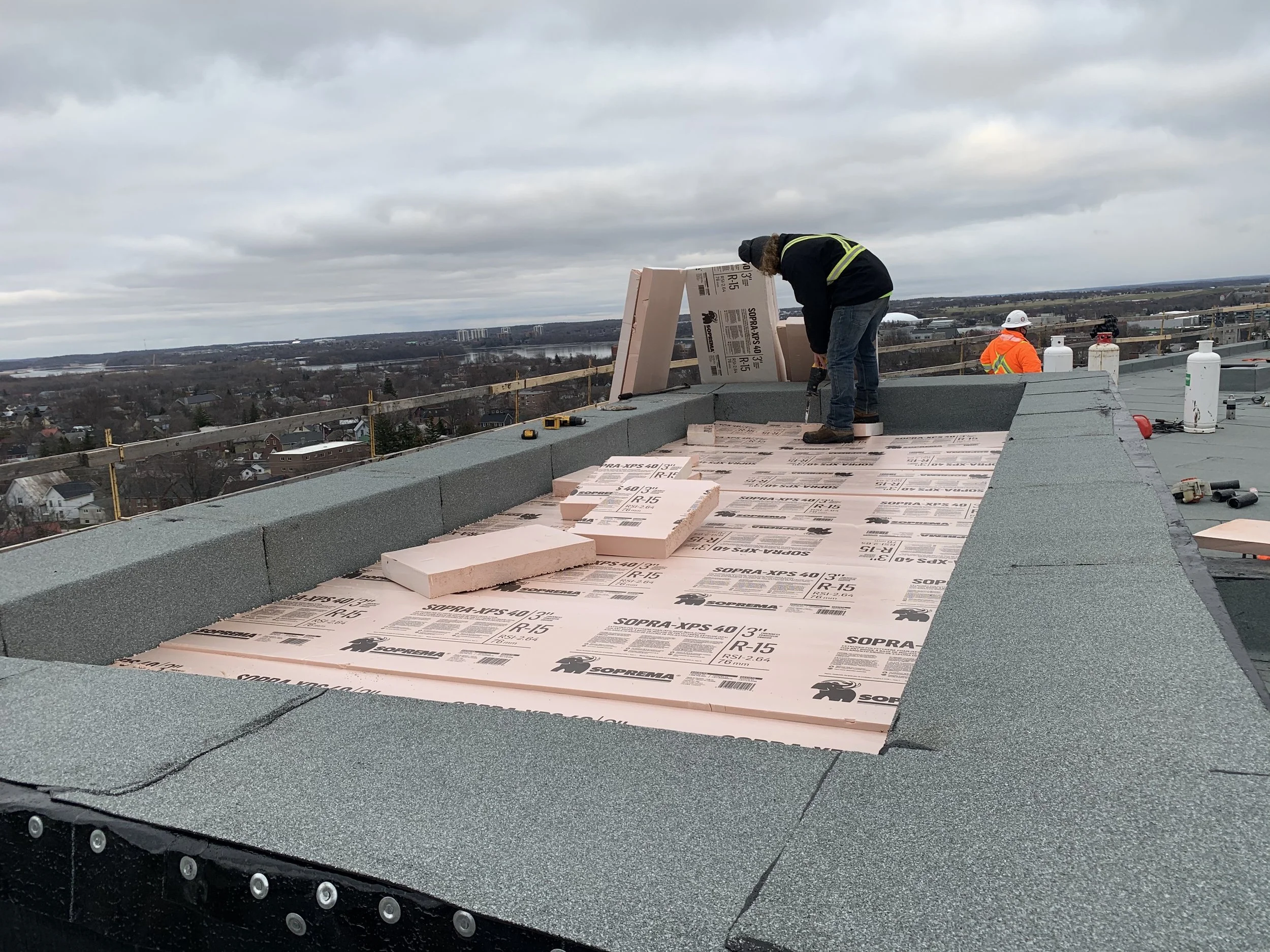 Construction workers on a rooftop installing insulation panels, with residential and urban landscape in the background under an overcast sky.