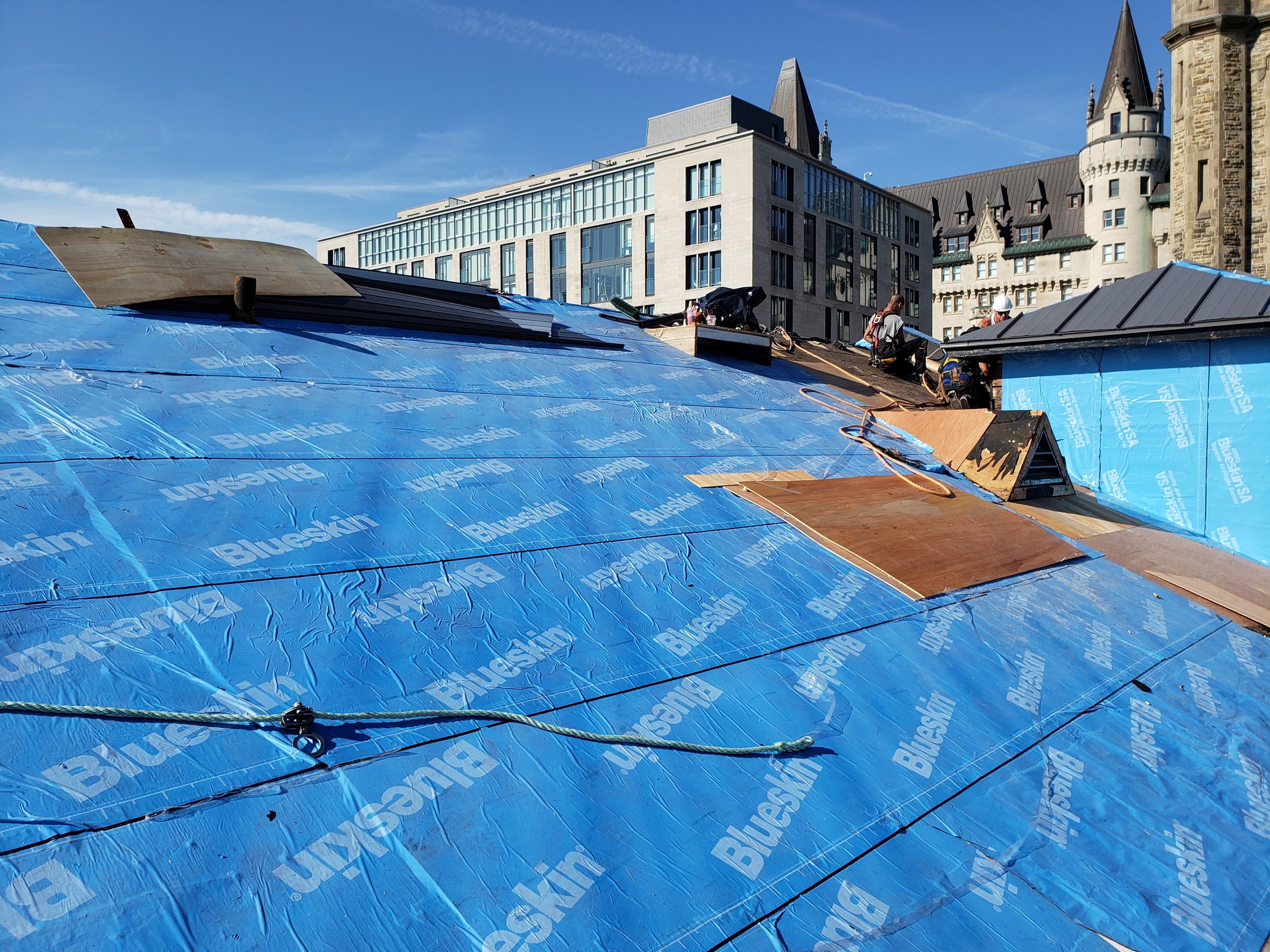 Construction workers installing blue roofing underlayment on a building in an urban downtown area, with modern and historic buildings visible in the background.