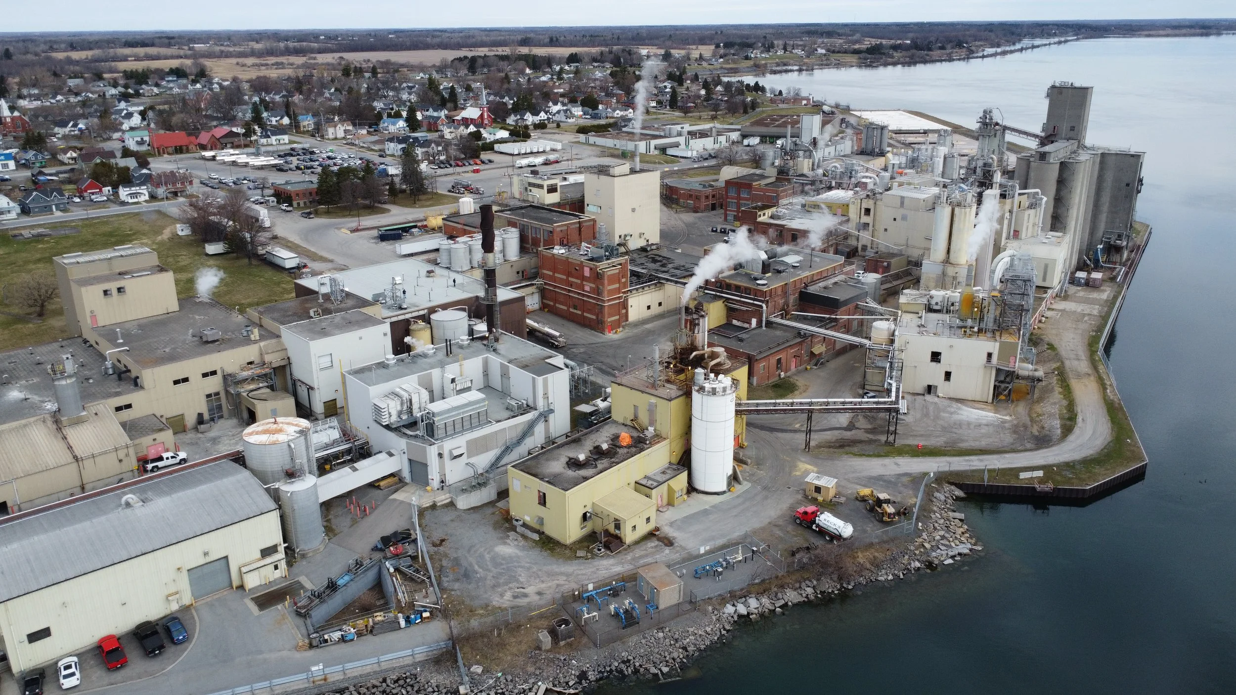 Aerial view of an industrial factory complex near a body of water, with a residential neighborhood in the background.
