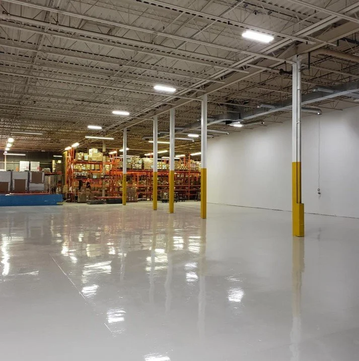 Empty warehouse with white painted shiny floor, metal shelving units with boxes in the background, white walls, and ceiling with fluorescent lights.