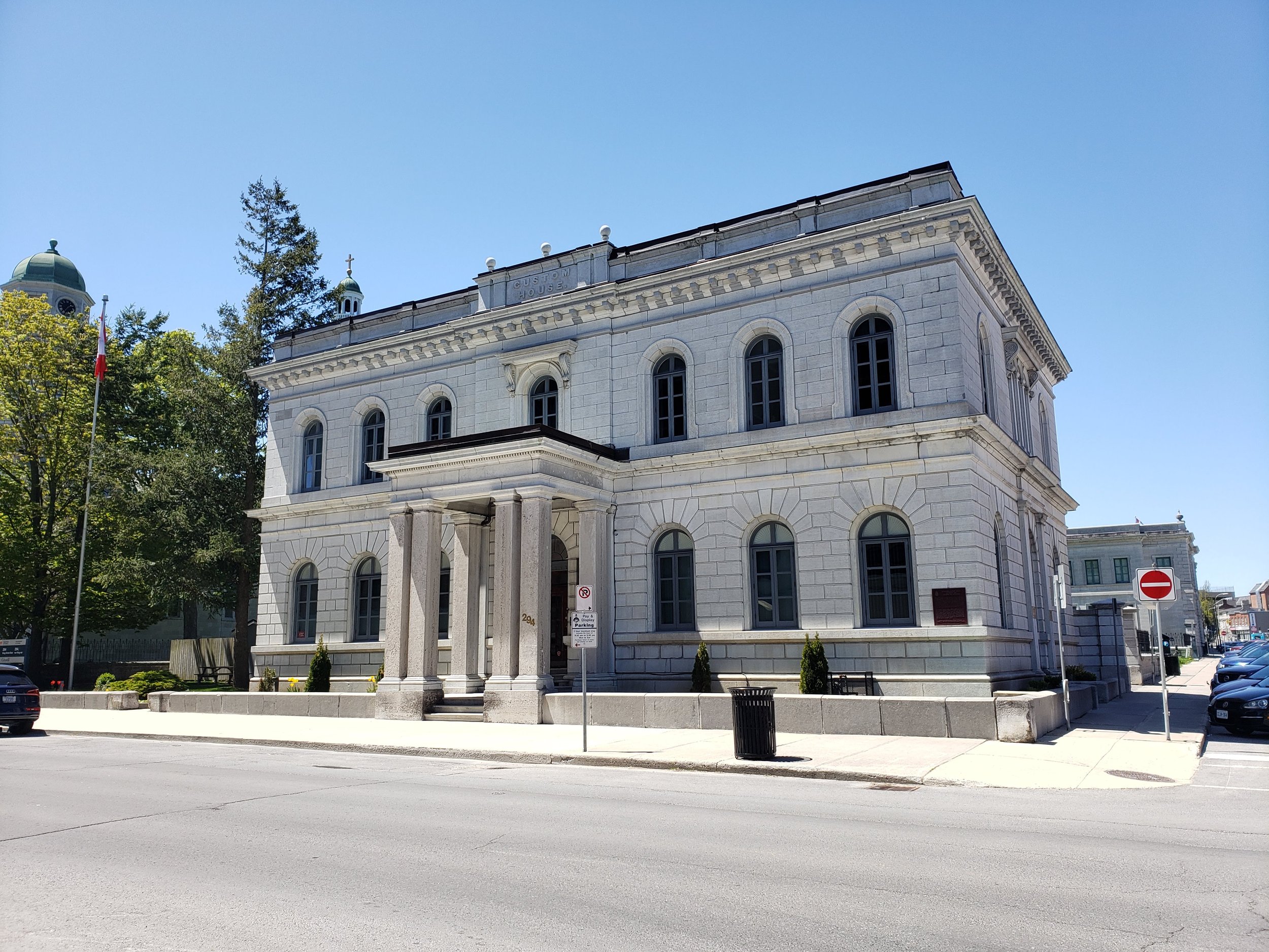 A historic white stone courthouse with tall windows and columns at the entrance, situated on a city street with cars parked nearby, green trees, a flagpole with the Canadian flag, and a clear blue sky.