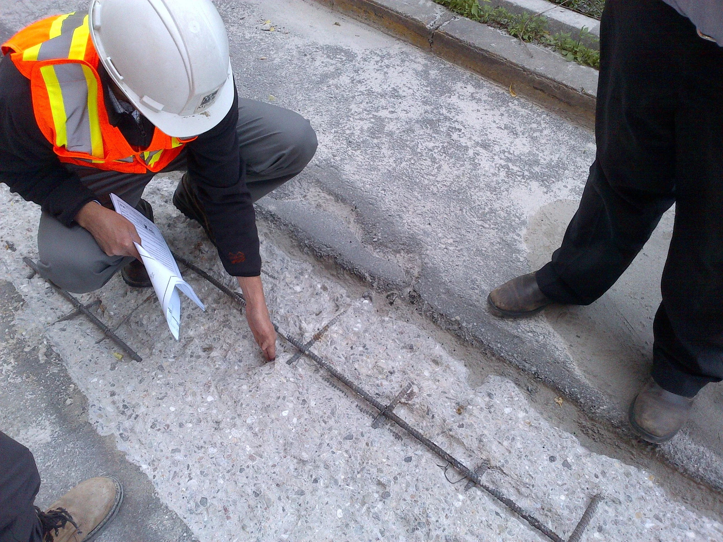 Construction workers studying a concrete curb or edge on a sidewalk, with one worker pointing at the ground and holding a clipboard, wearing a white safety helmet and a bright orange safety vest. The scene is outdoors on a sidewalk with some grass and gravel.