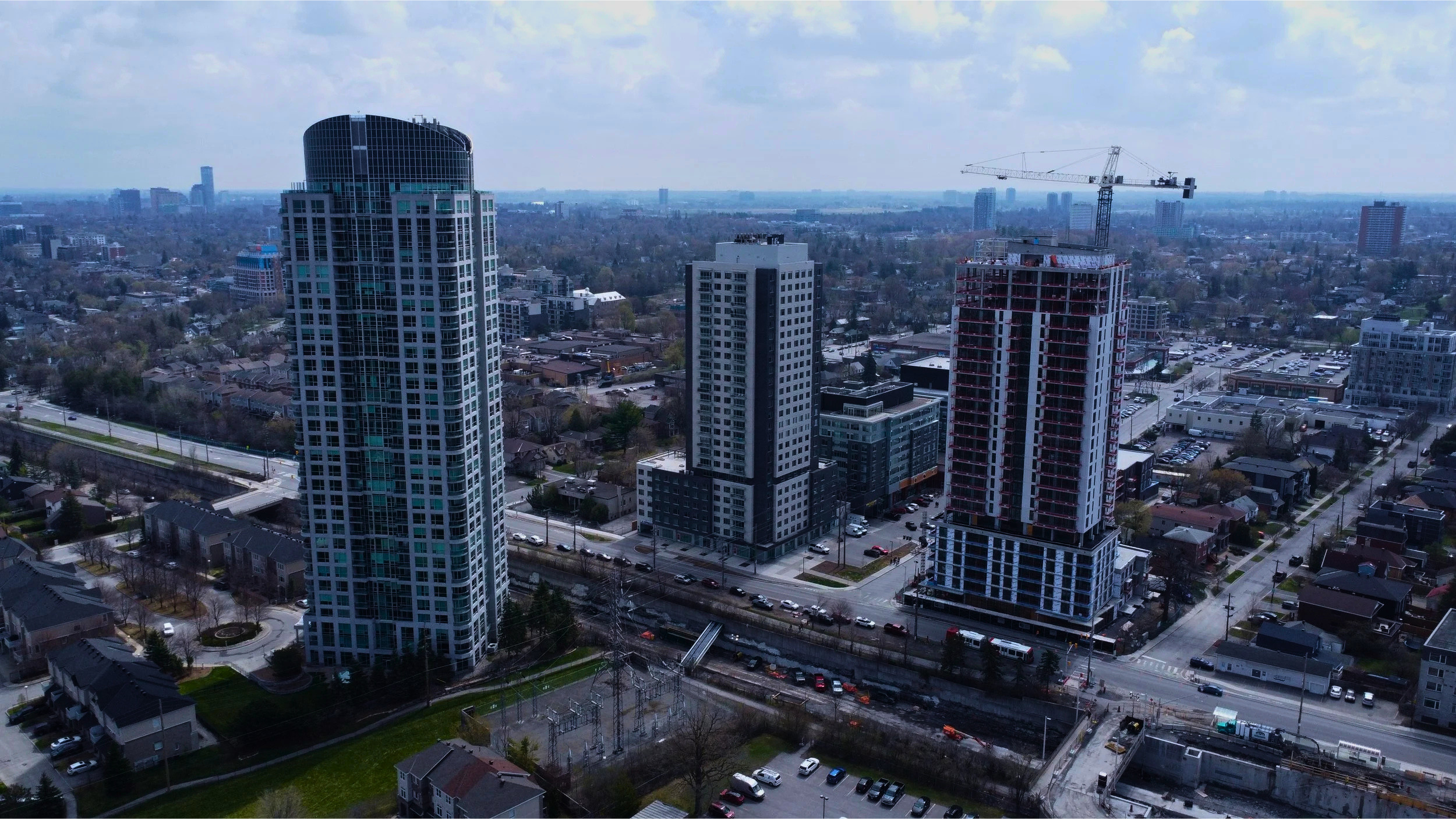 Aerial view of a cityscape with tall buildings, residential houses, and streets, with a distant skyline and a construction crane adding to the urban environment.