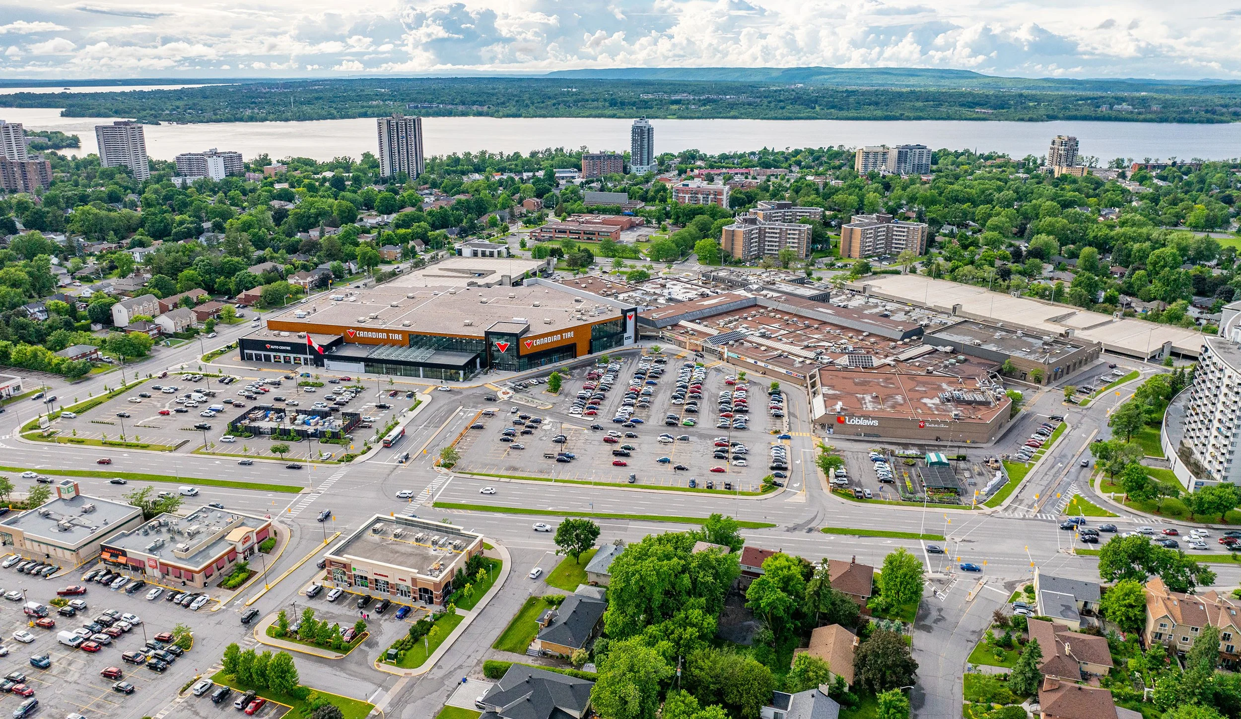 Aerial view of a city with a shopping mall surrounded by parking lots, residential houses, mid-rise buildings, and a river in the background under a partly cloudy sky.