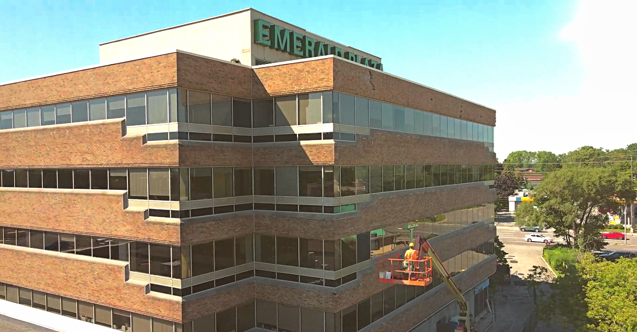 A multi-story brick office building with large windows, with a worker on an elevated platform cleaning the windows. The building has Green text on top, partially visible, and is located in a suburban area with trees and cars in the parking lot.