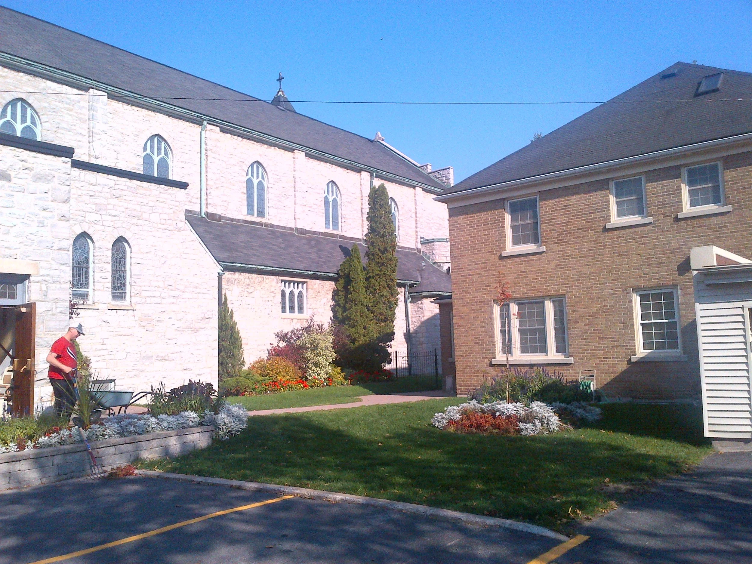 A man in a red shirt and straw hat watering flowers in a garden with a wheelbarrow nearby, in front of a stone and brick church with arched windows and a steeple, blue sky overhead.