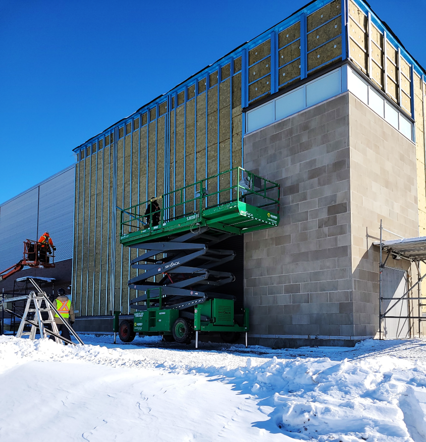 Construction workers install insulation on an exterior building wall using a green scissor lift, with snow on the ground and additional construction equipment nearby.