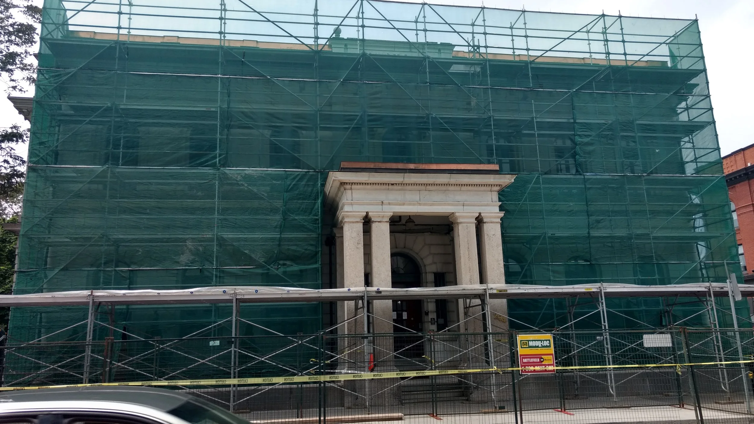 A building under renovation covered by green construction netting and scaffolding. The building has a classical entrance with columns and an arched window.