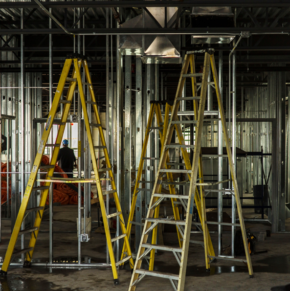 Interior of a building under construction with multiple yellow and silver ladders, metal framing, and construction workers in the background.