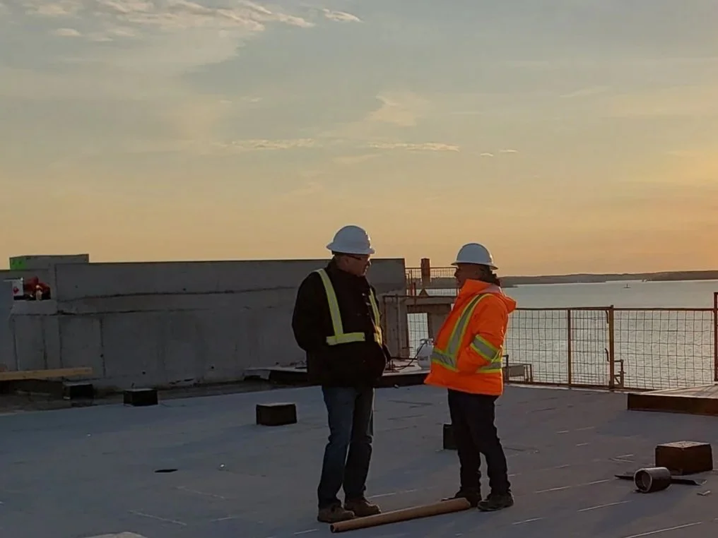 Two construction workers wearing hard hats and safety vests standing on a building rooftop, discussing while holding a clipboard, with a body of water and sunset in the background.