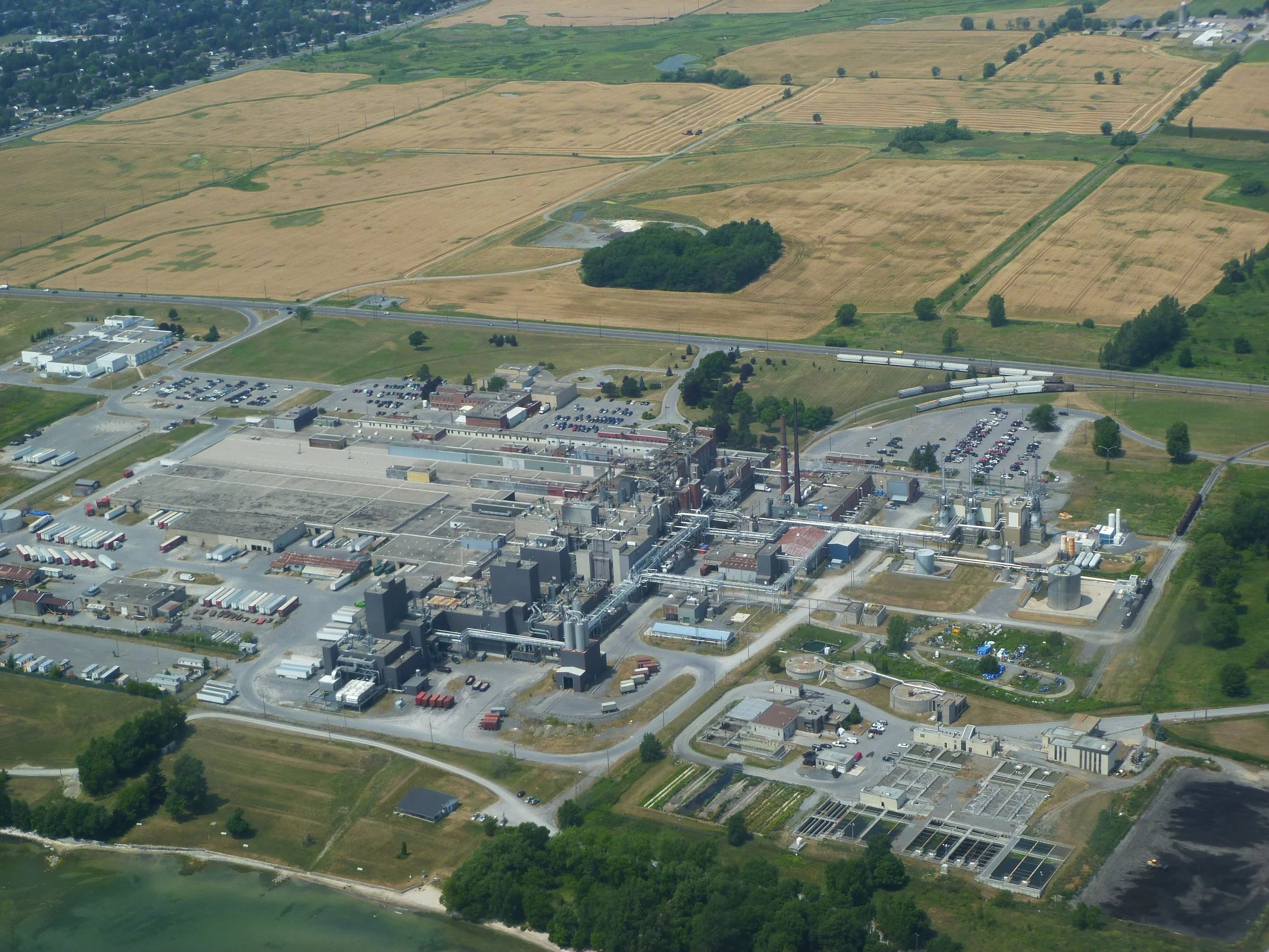 An aerial view of a landscape showing a mix of agricultural fields, a factory or industrial complex, and a body of water. The landscape includes patches of green trees, roads, and a parking lot.
