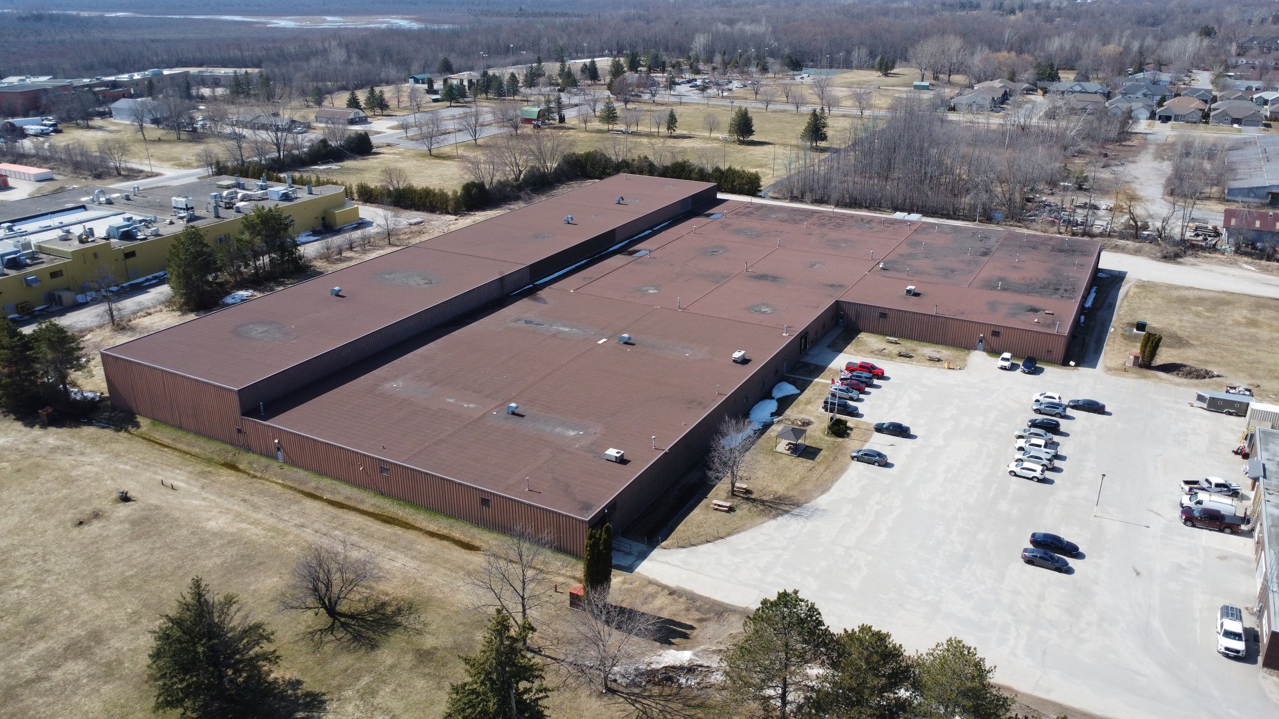 An aerial view of a large building with a brown roof, a parking lot with several cars, and surrounding green open spaces and trees.
