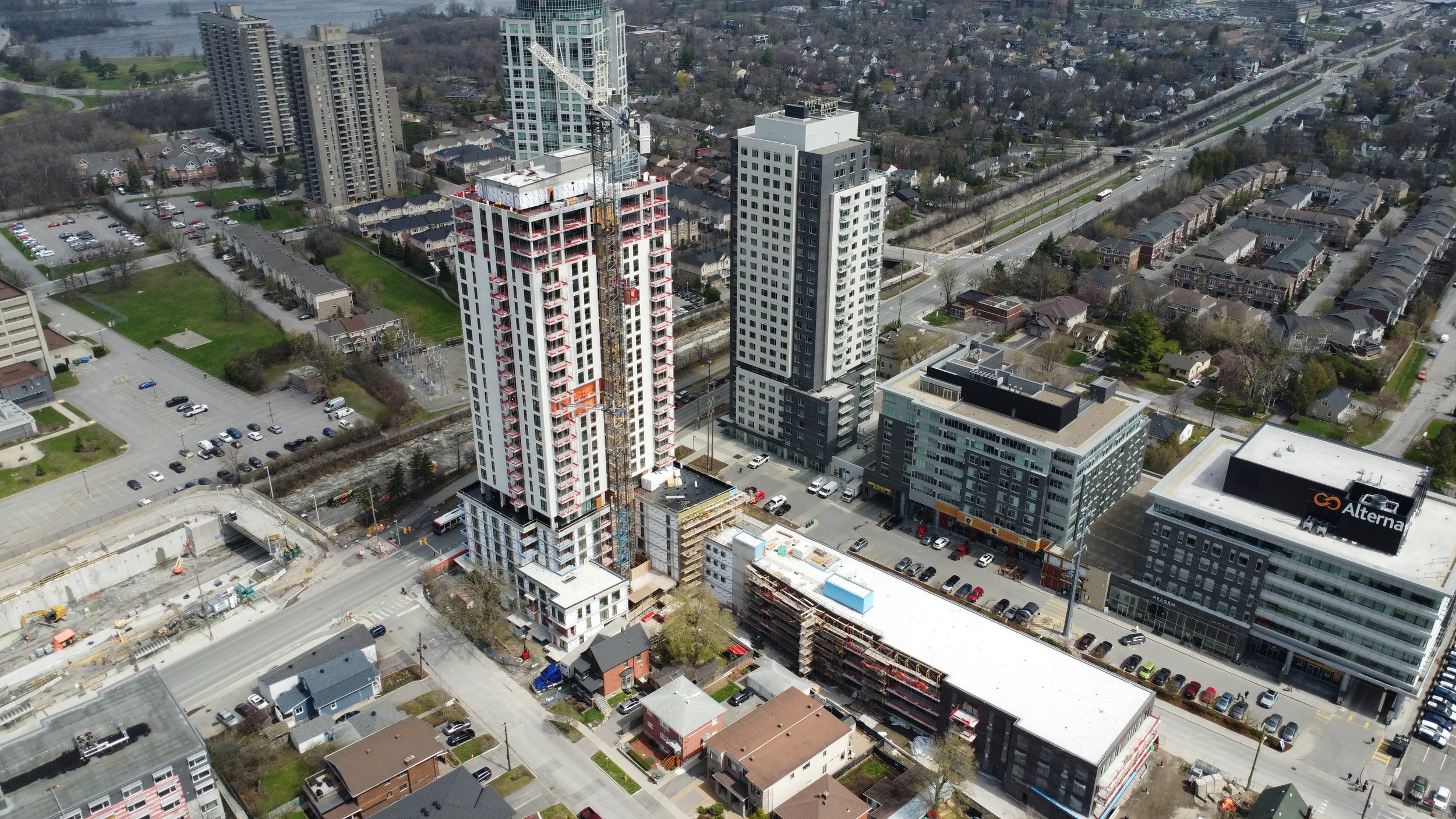An aerial view of a cityscape showing high-rise buildings under construction, surrounding residential houses, a parking lot, and a highway in the background.