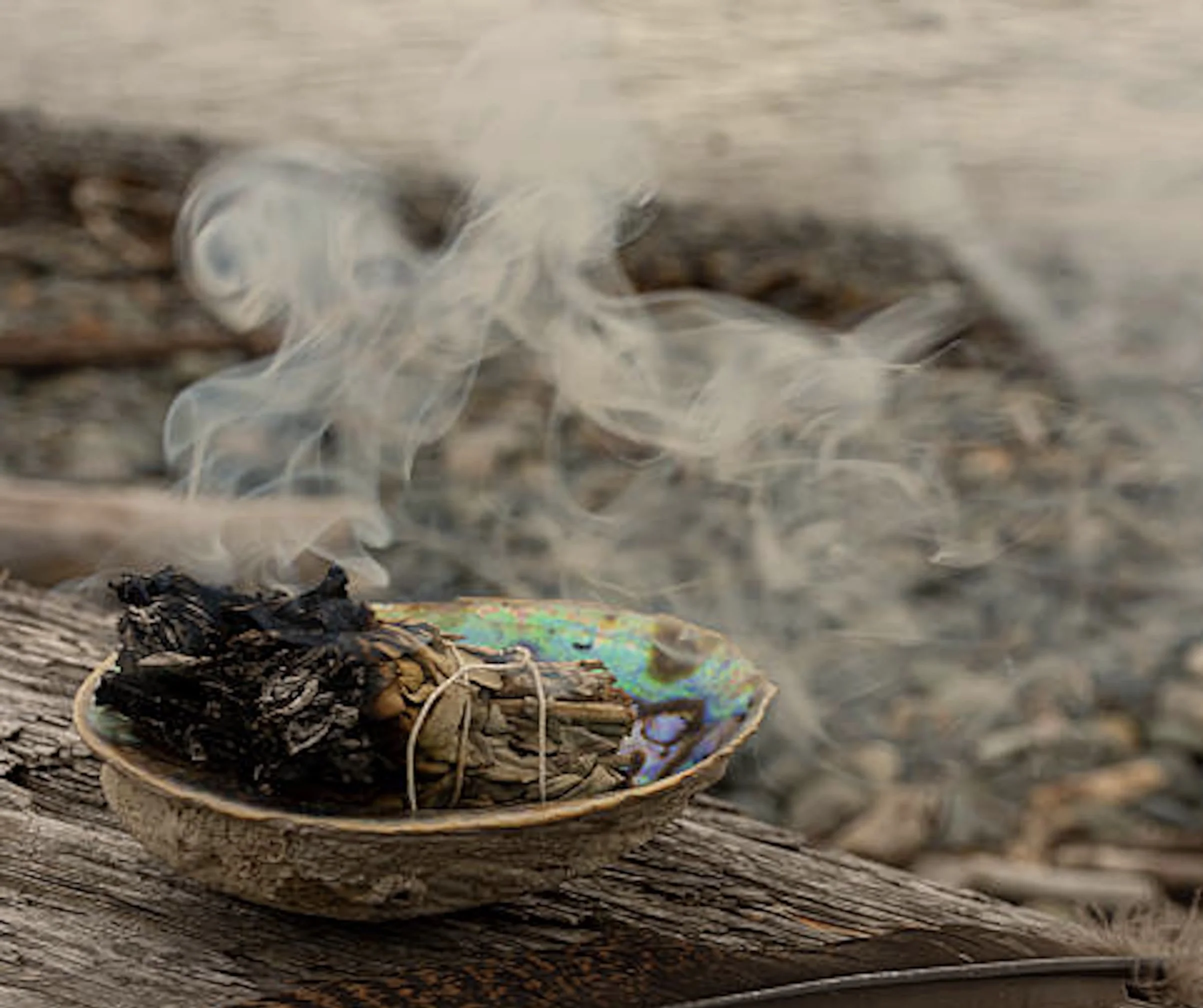 A beautifully focused shot of a ceramic smudge dish holding a bundle of white sage next to loose ritual tobacco leaves and a box of wooden matches on a dark table.