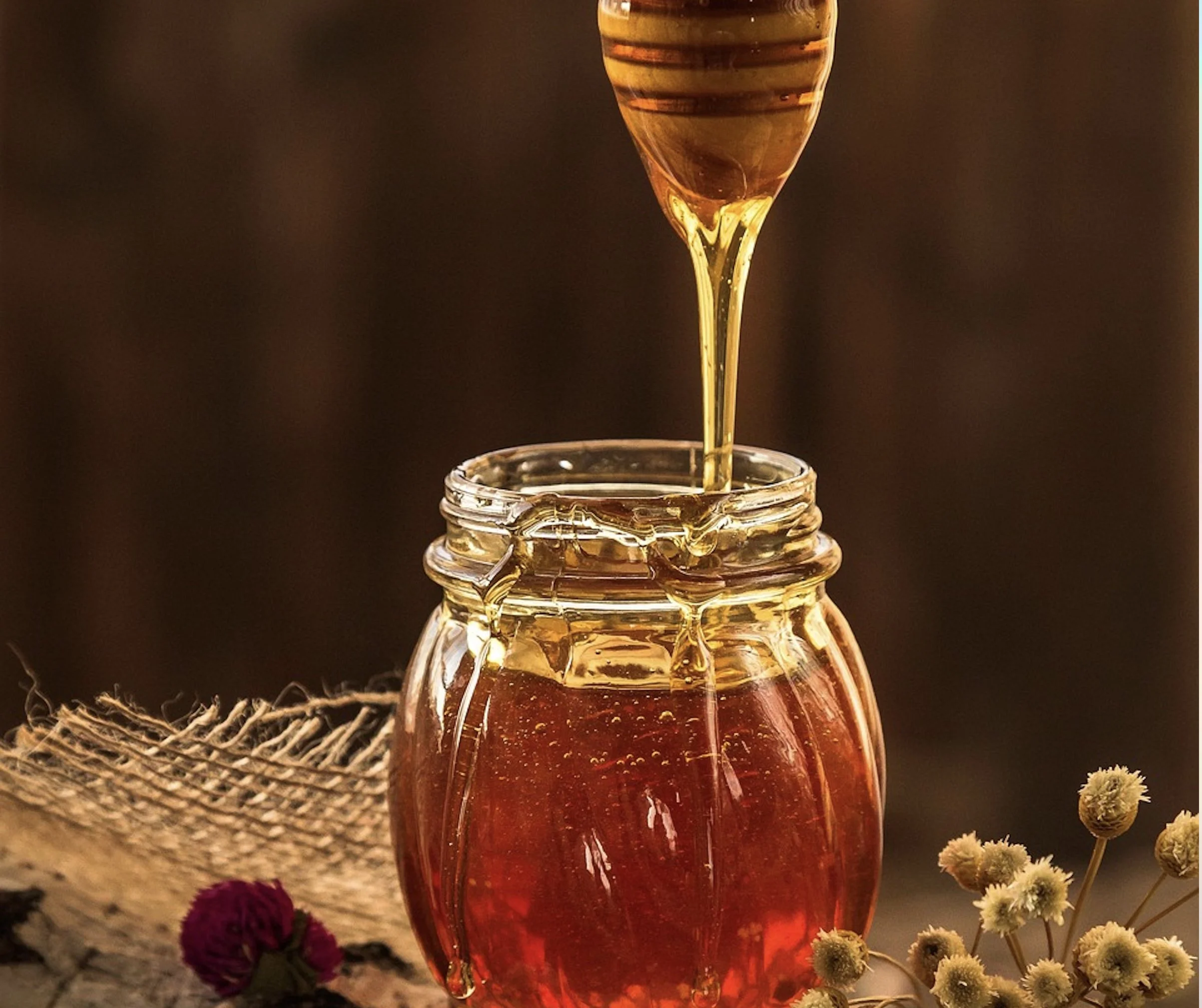 A beautifully warm close-up of a small glass jar filled with golden honey, resting on a dark cloth next to loose pink rose petals and a burning pink candle.