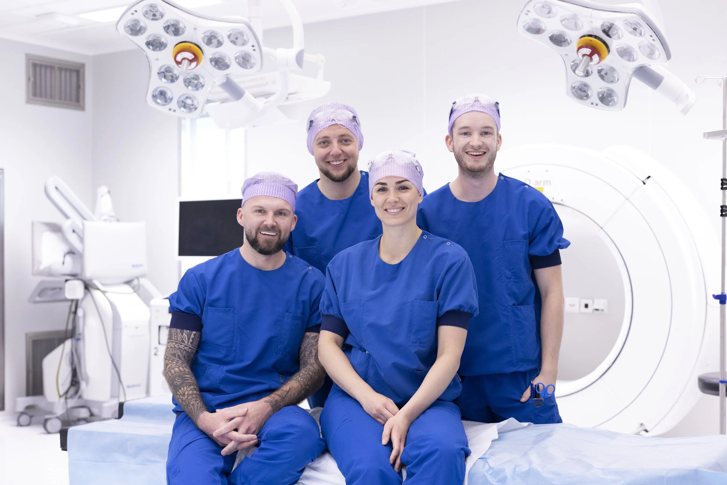 Vijf medisch gekleedde professionals zittend en staand in een operatiekamer met medische apparatuur en verlichte chirurgielampen op de achtergrond.