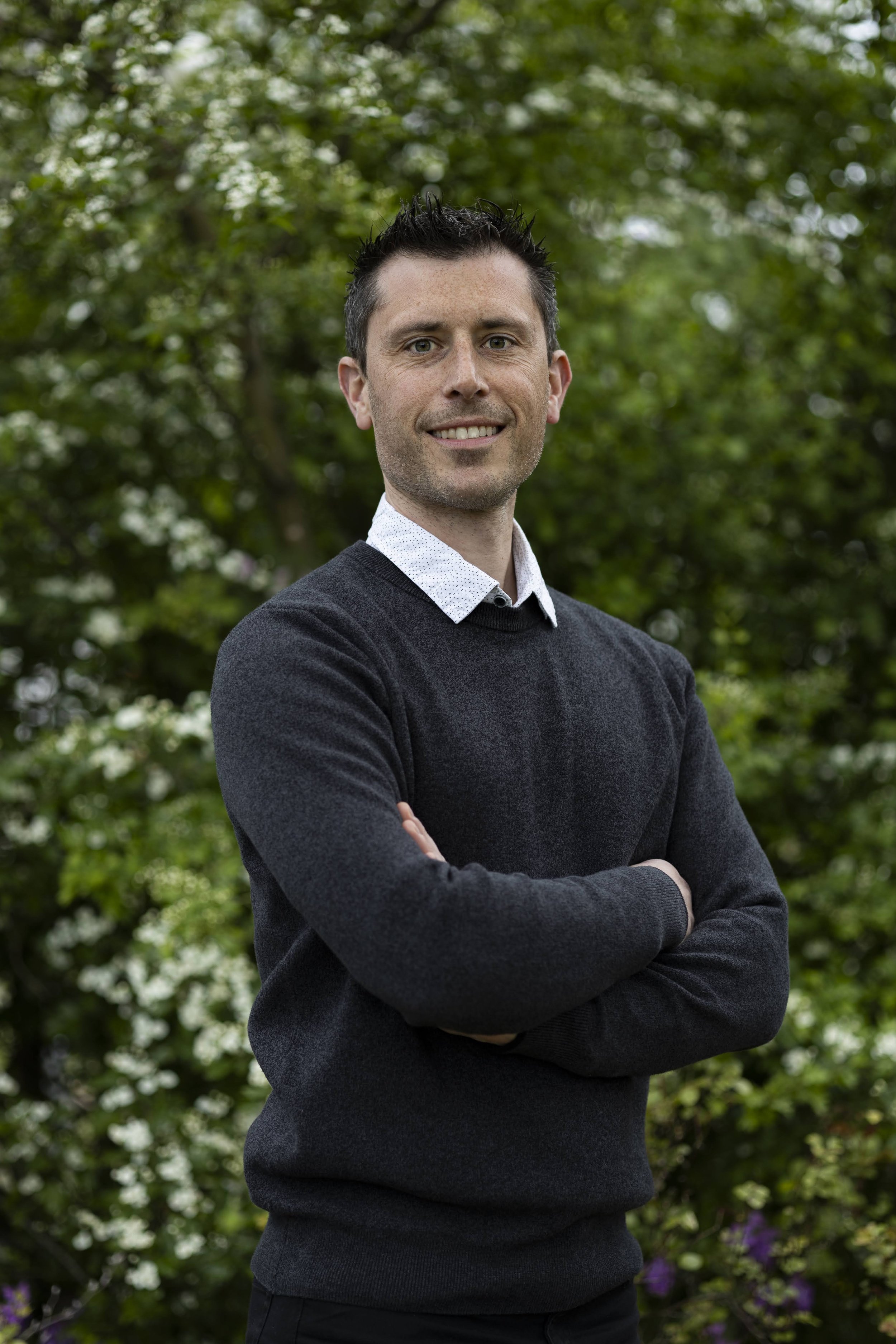 Man met korte donkere haren en een lichte huid, lachend, gekleed in een wit overhemd en een donkergrijze trui, staand met armen over elkaar voor een groene, bloeiende achtergrond.