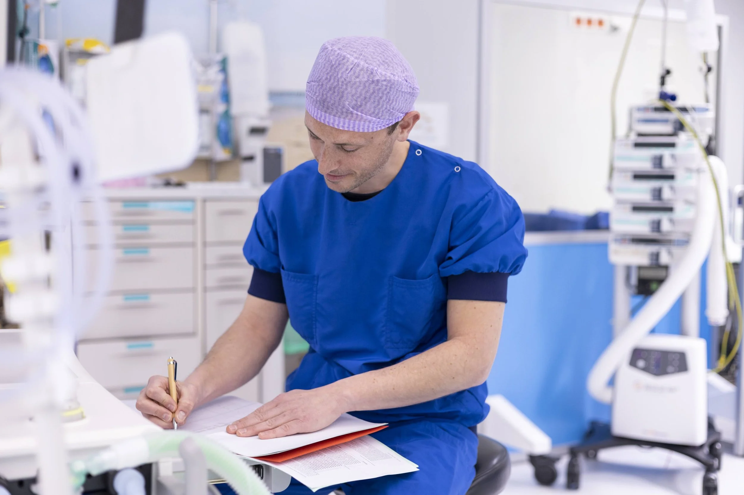 een man in blauwe medische kleding en een paarse hoofddeksel die onder een tafel schrijft in een klinisch laboratorium.