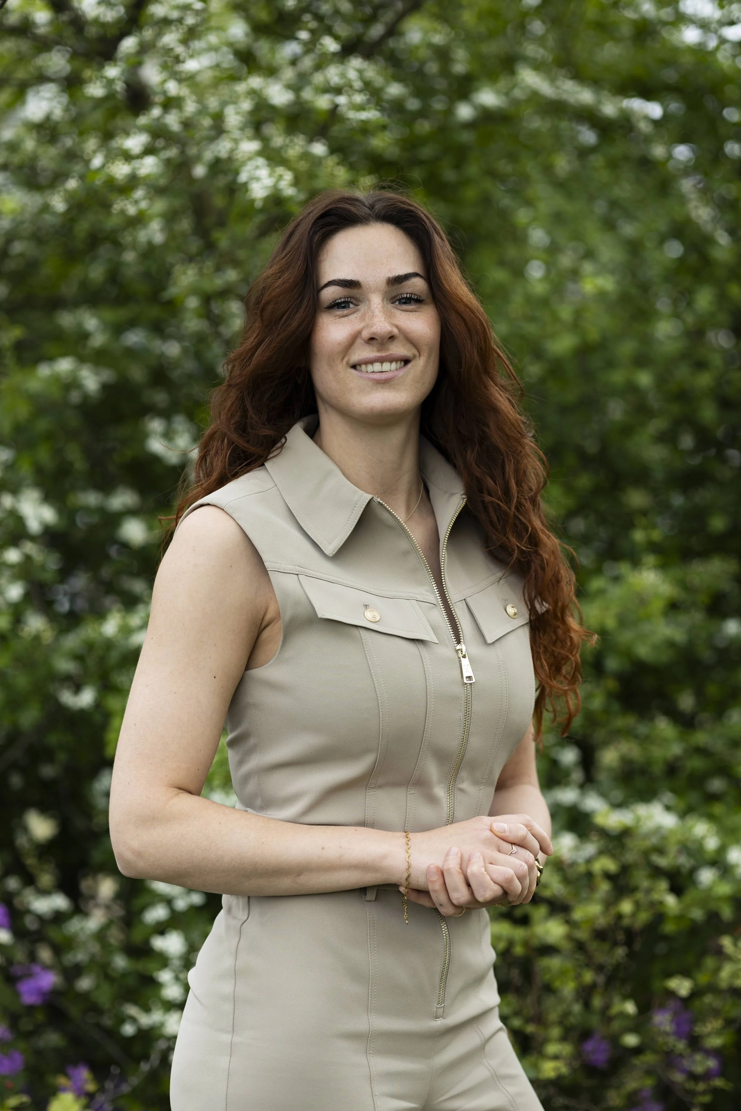 Portret van een jonge vrouw met lang, kastanjebruin haar en een lichte beige jurk, glimlachend, buiten met groene bladeren op de achtergrond.
