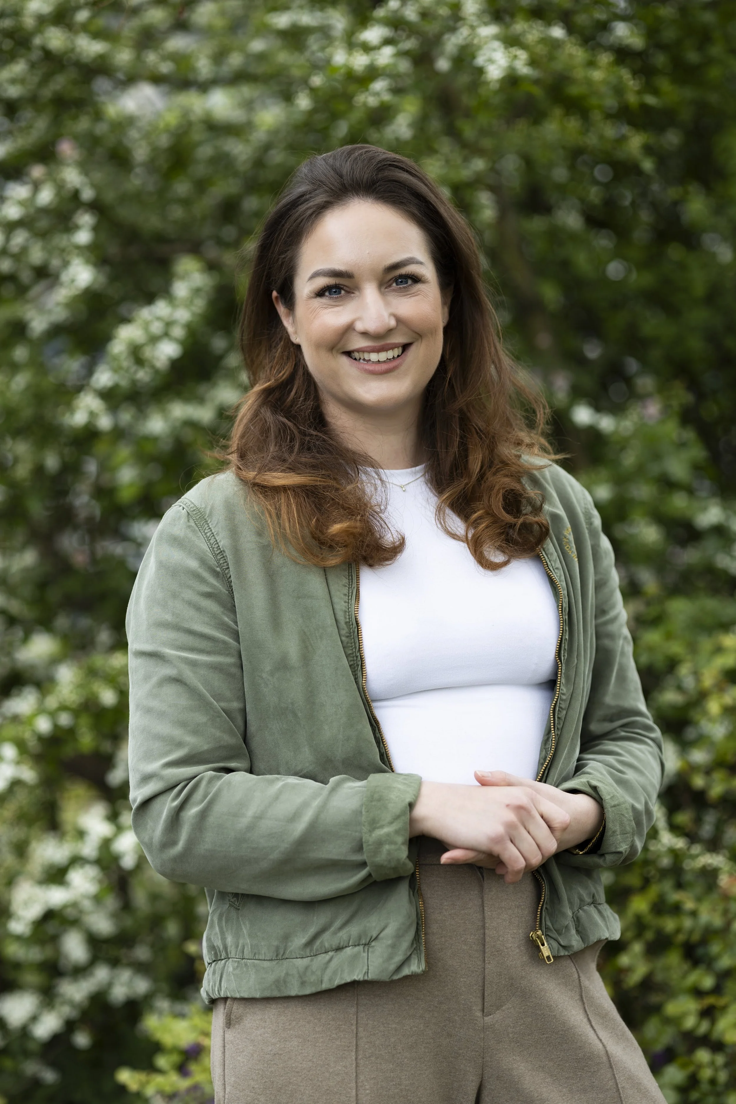 Een lachende jonge vrouw met bruin haar, gekleed in een witte T-shirt en een groene jas, staand in een tuin met groene bladeren op de achtergrond.