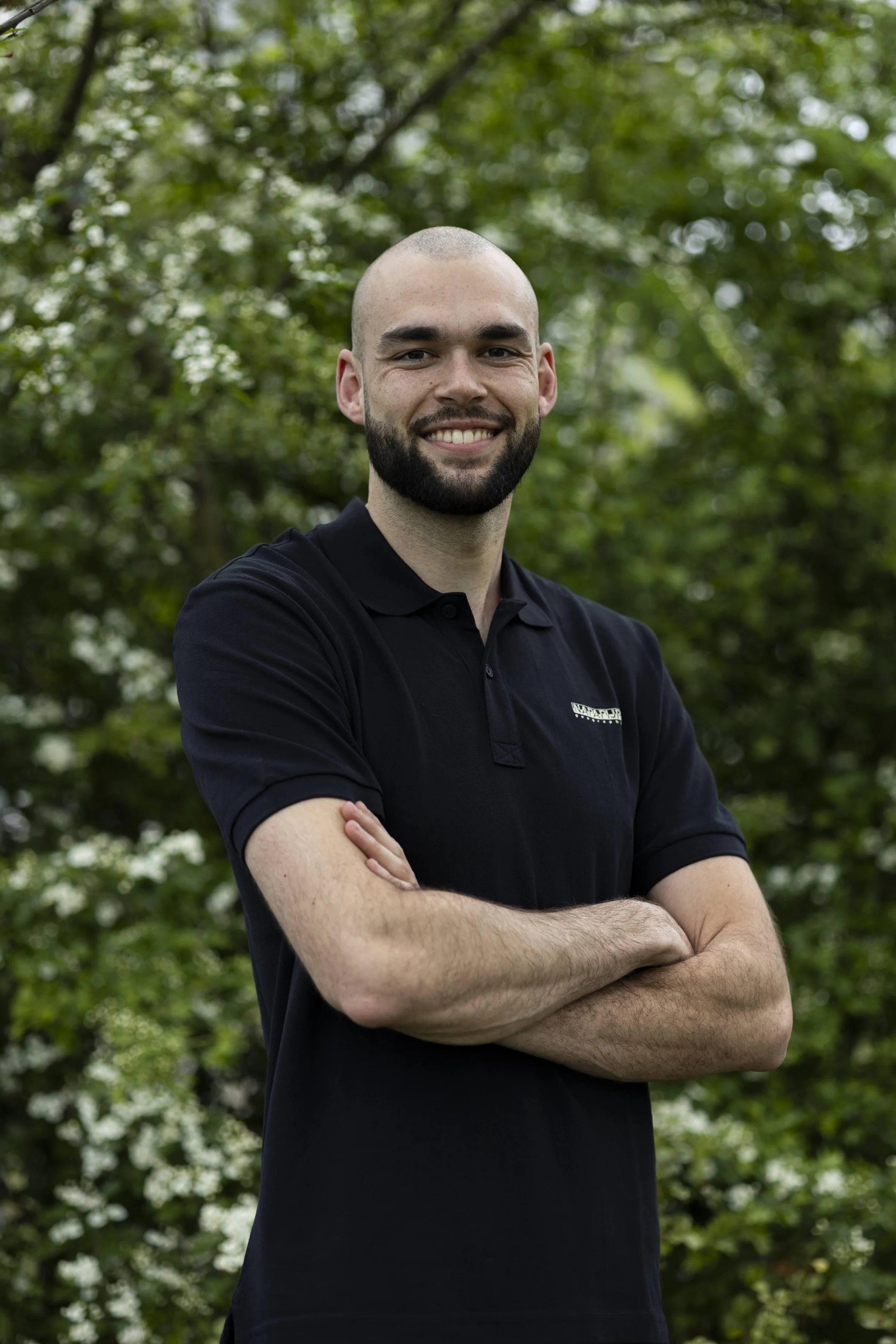 Man met ontbloot hoofd, glimlachend, gekleed in een zwart polo, met zijn armen gekruist, buiten met groene bomen op de achtergrond.