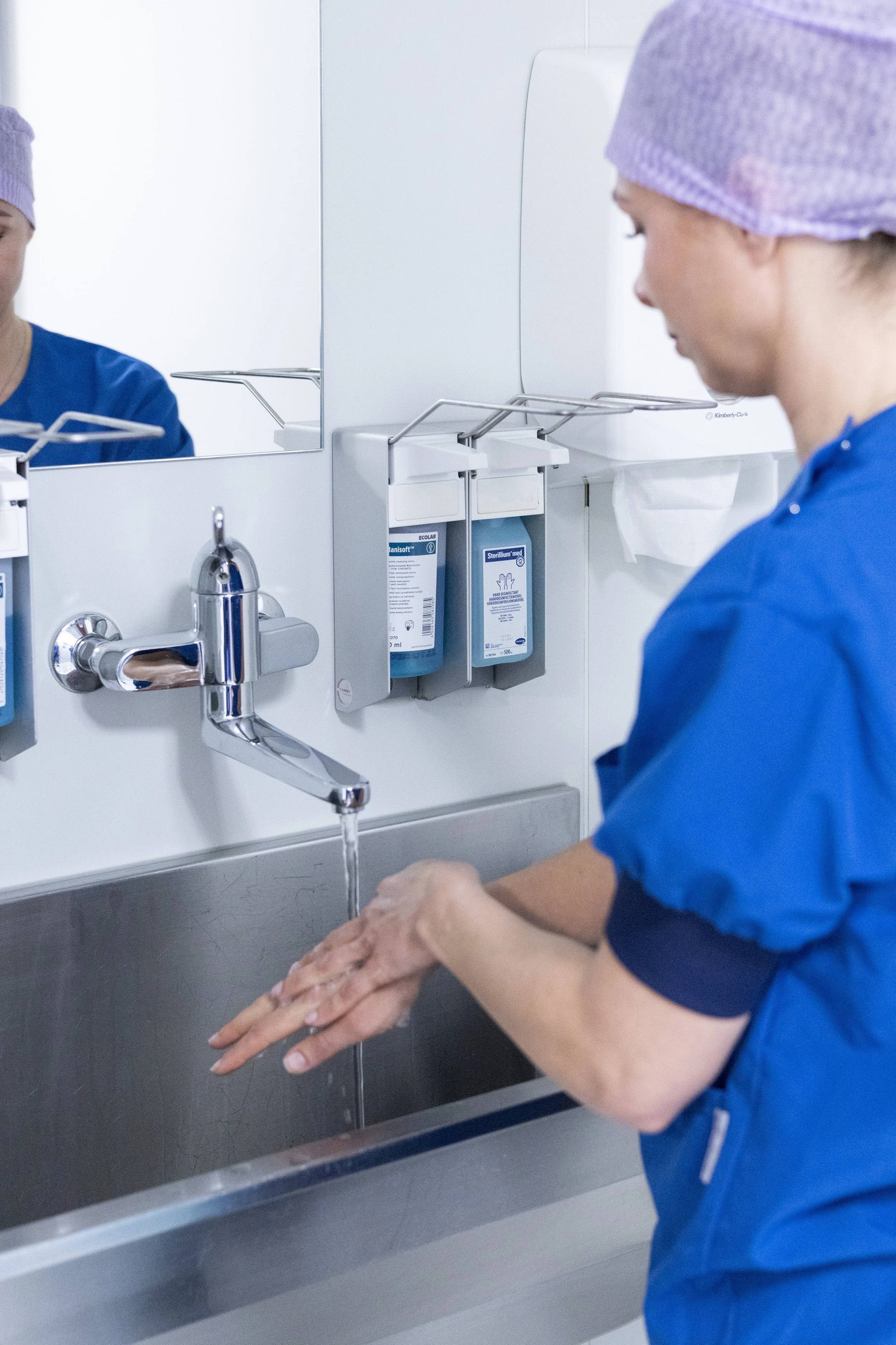 Een vrouw wast haar handen onder een lopende kraan in een medische of laboratoriumomgeving, gekleed in blauwe medische uniformen en paarse haarpetjes.