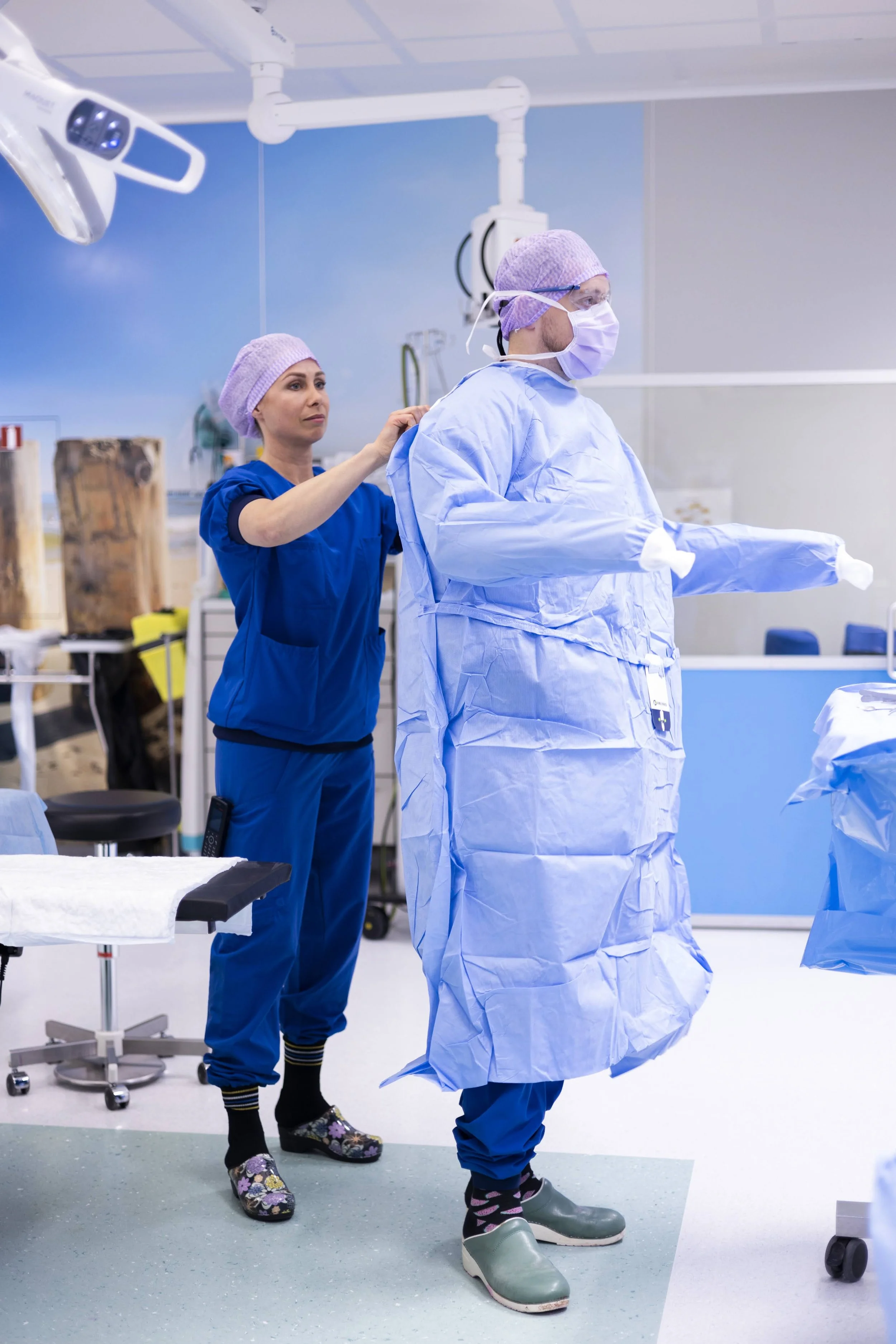 Twee medisch personeel in een operatiekamer. Eén persoon helpt een andere die in een operatiepak staat, allemaal dragen ze maskers, mutsen en beschermende schoenen.
