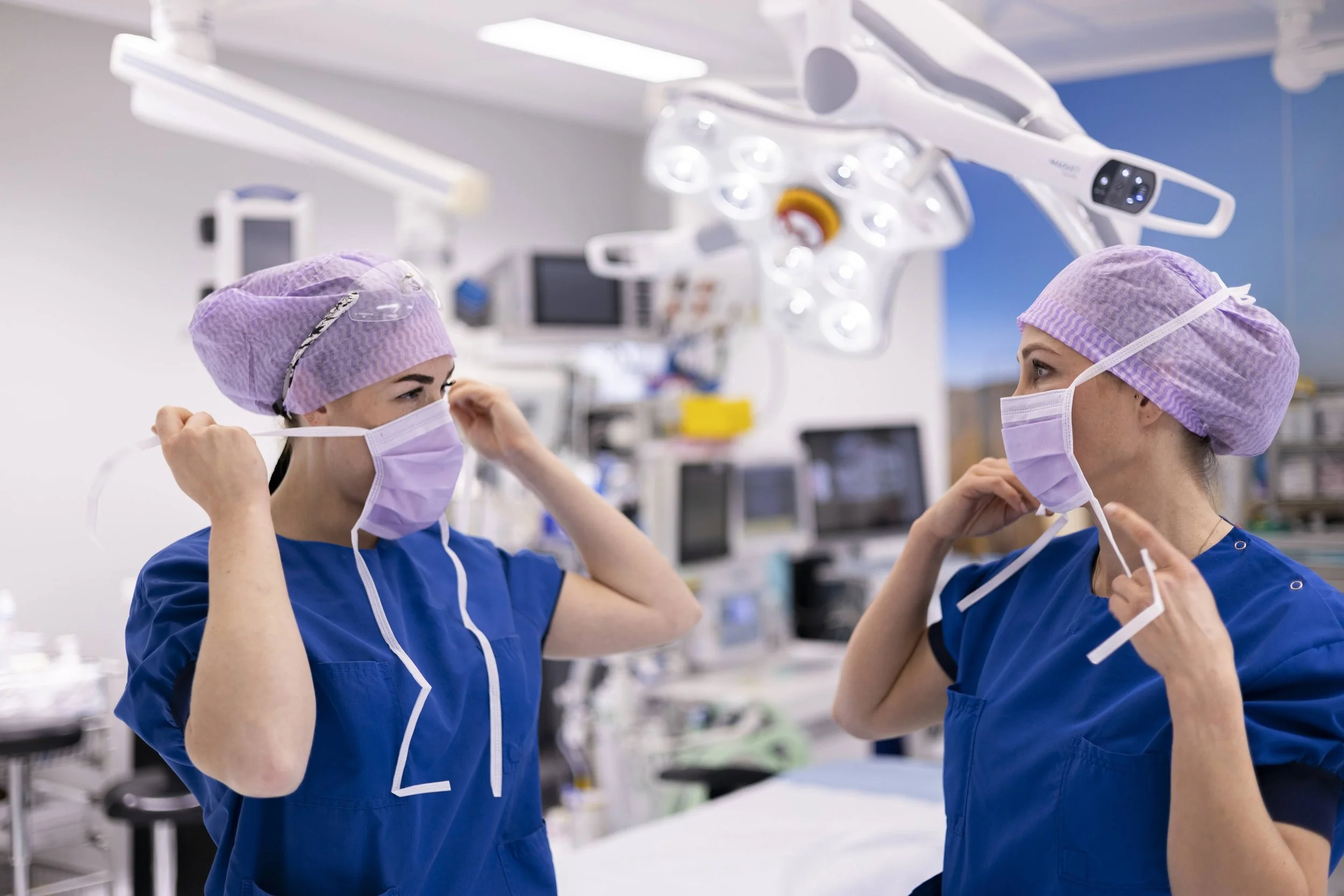 Twee medische professionals dragen blauwe uniformen, paarse haarkappen en gezichtsmaskers in een operatieruimte, die om zich heen medische apparatuur bevat.