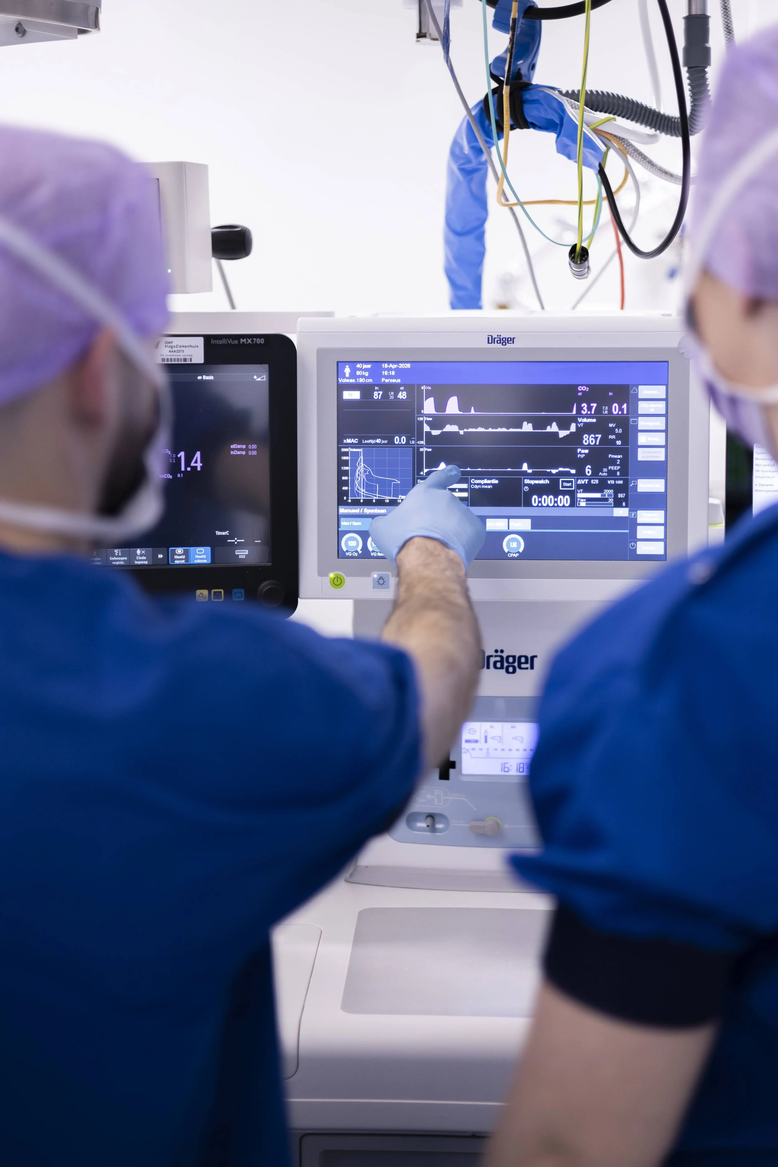 Medisch personeel bedient een beademingsmachine in een ziekenhuis. Ze dragen blauwe uniformen en handschoenen, en er hangen kabels en medische apparatuur boven de machine.