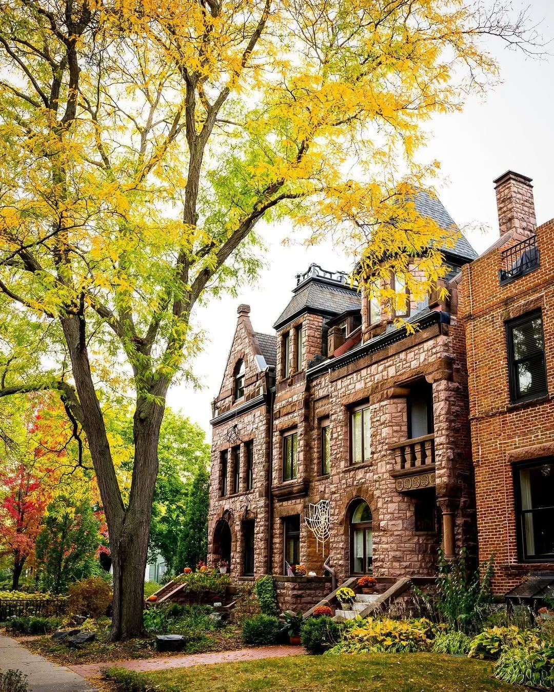 A historic brick house on Summit Avenue with a turret, decorative stonework, and a spider web on the front steps, surrounded by colorful fall trees with yellow and red leaves.