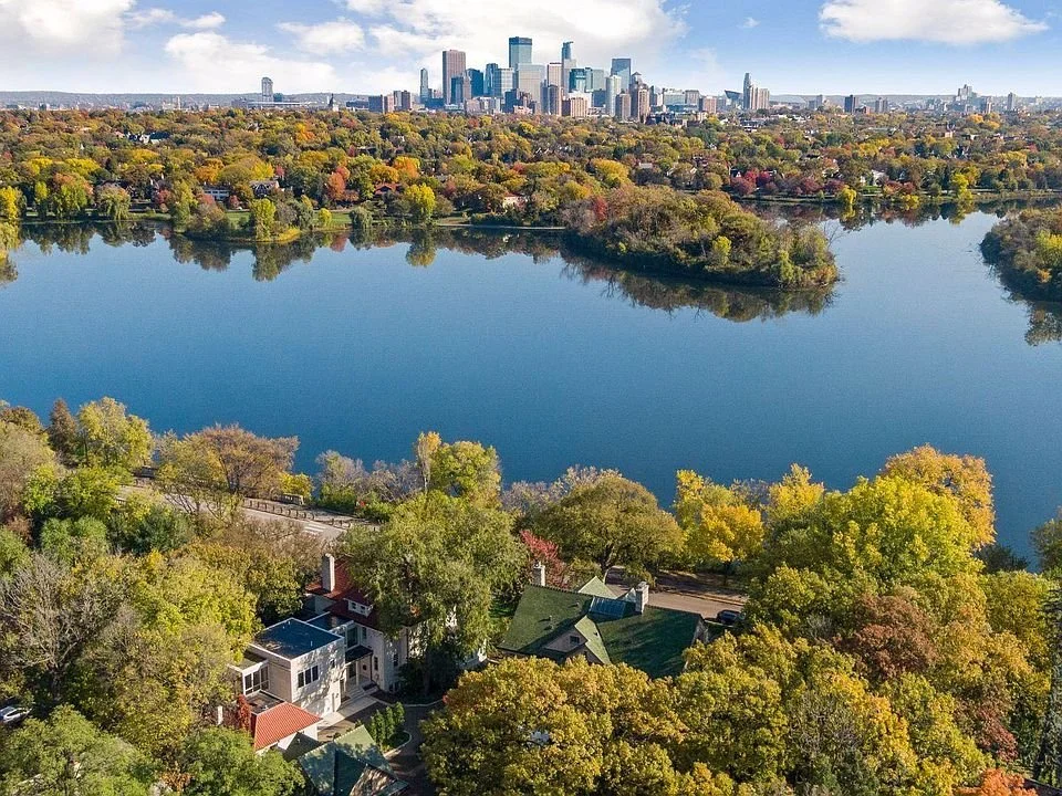 Aerial view of a lake with trees in fall colors, residential houses in the foreground, and a city skyline in the background under a partly cloudy sky.