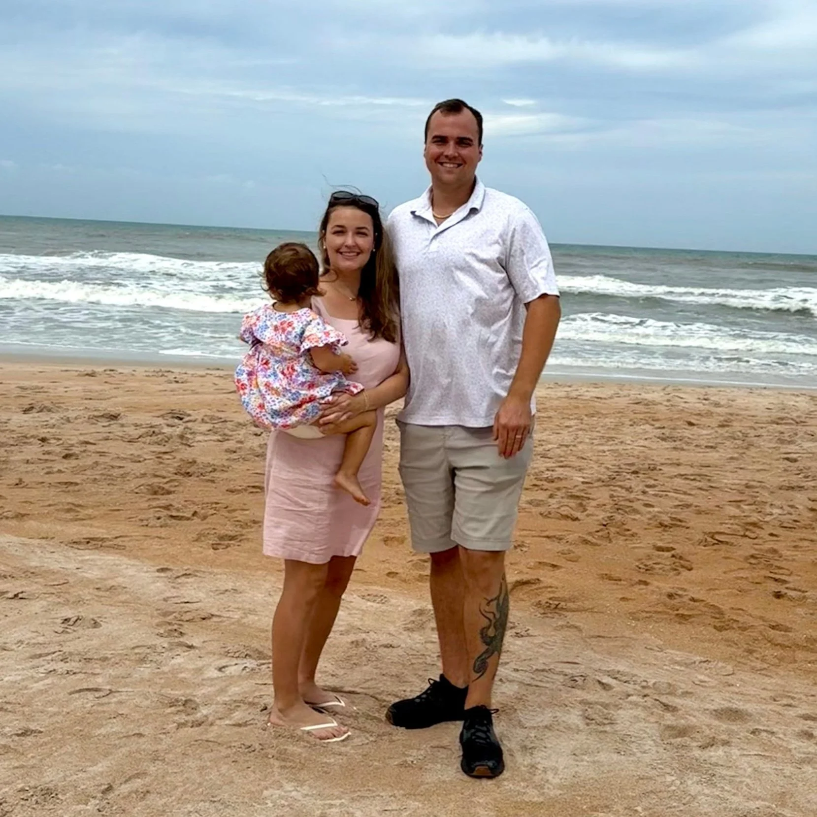 A family of three standing on a sandy beach with the ocean and cloudy sky in the background. A mother is holding a small child looking away from the camera, and her husband stands beside them smiling at the camera.