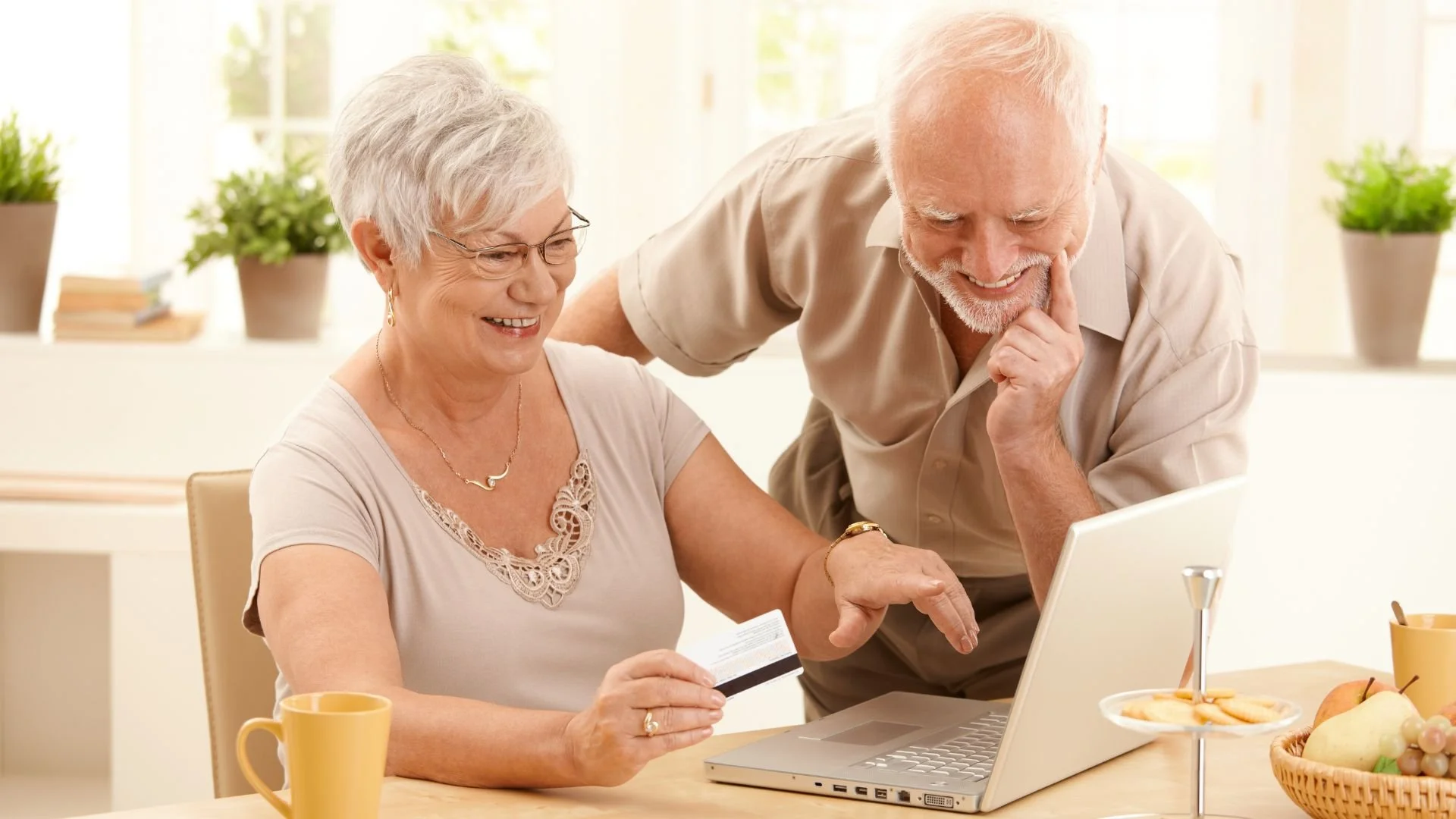 An elderly woman and man are sitting at a table, smiling and looking at a laptop screen. The woman is holding a credit card, and they appear to be shopping or banking online, with a bright, cheerful background featuring potted plants and a window.
