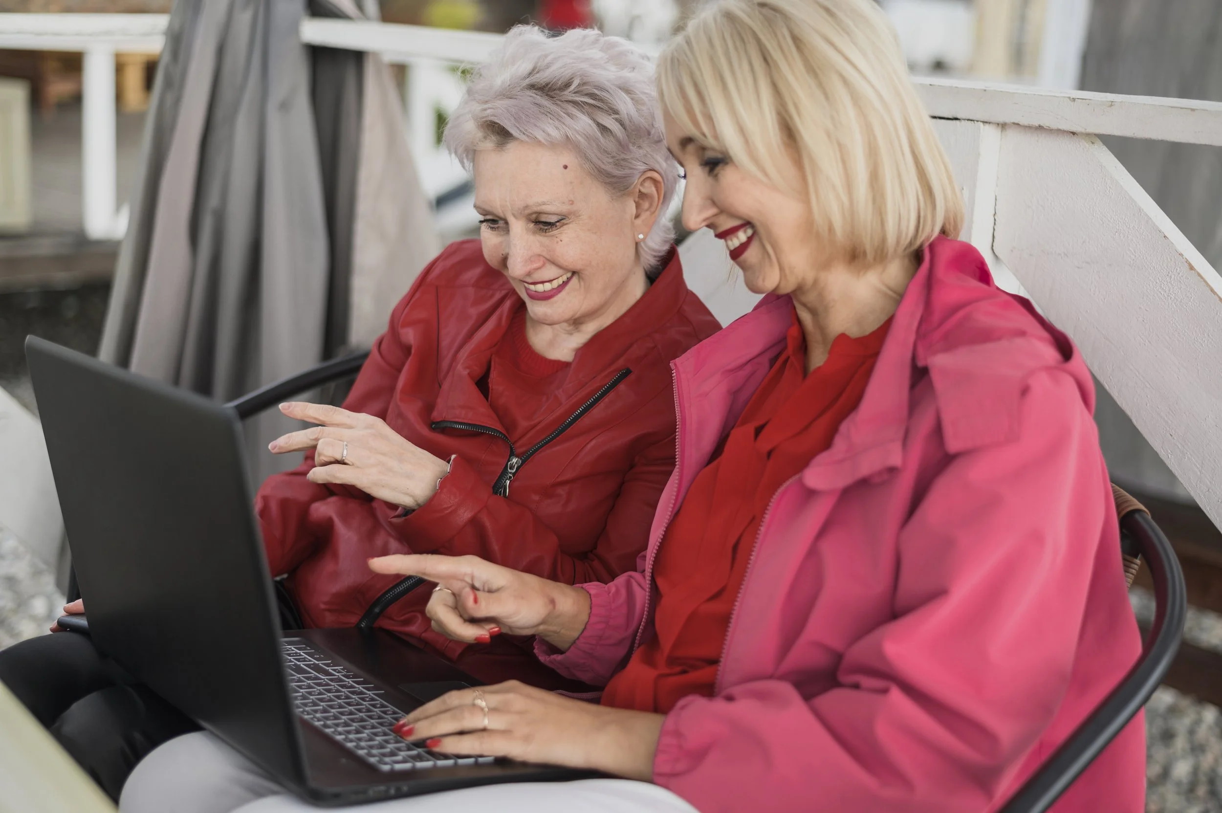 Two women sitting outdoors on a bench, looking at a laptop and smiling, enjoying a moment together.