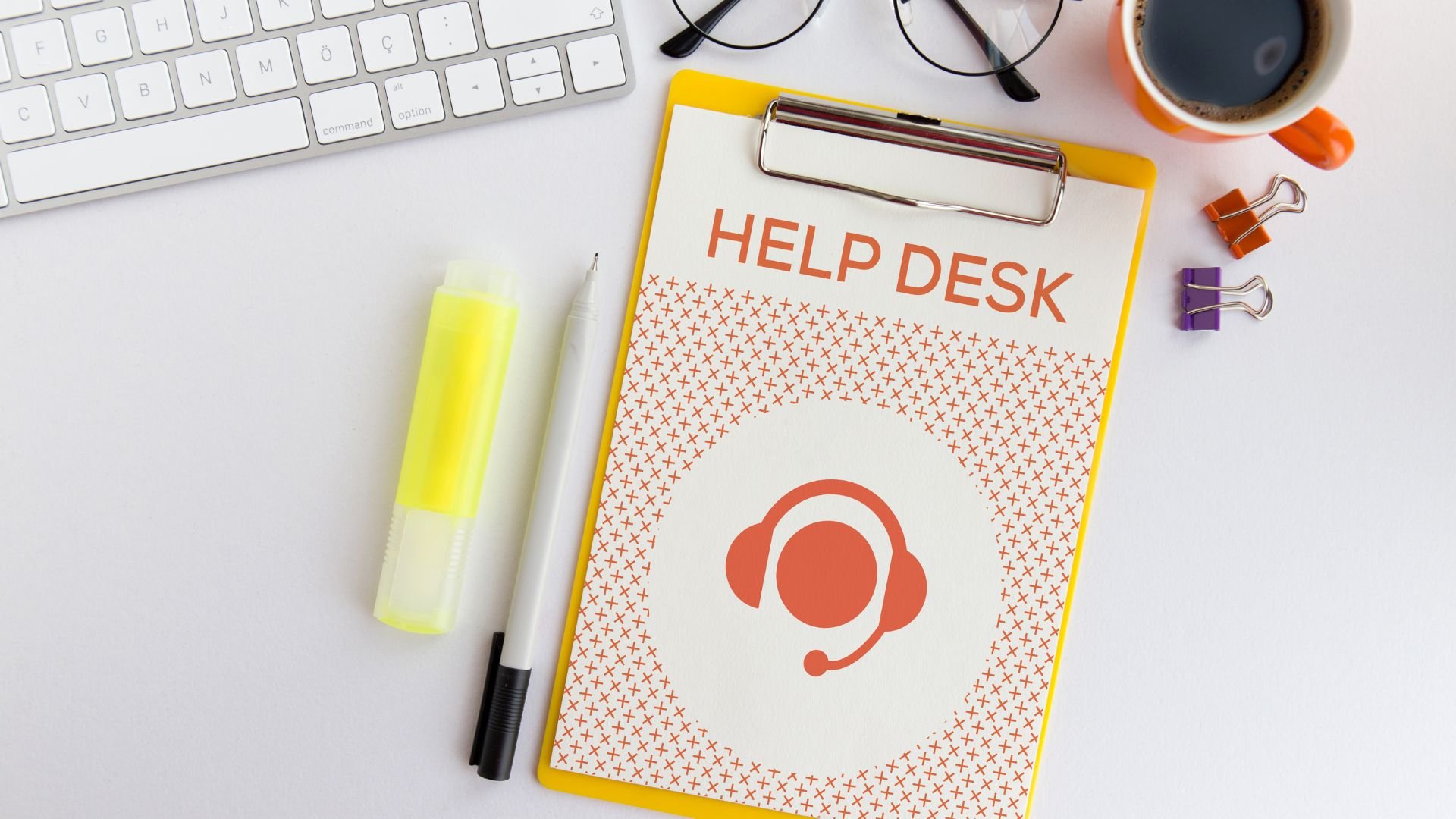 Desk with keyboard, clipboard labeled 'Help Desk', yellow highlighter, white pen, sunglasses, cup of coffee, and colorful binder clips on a white surface.