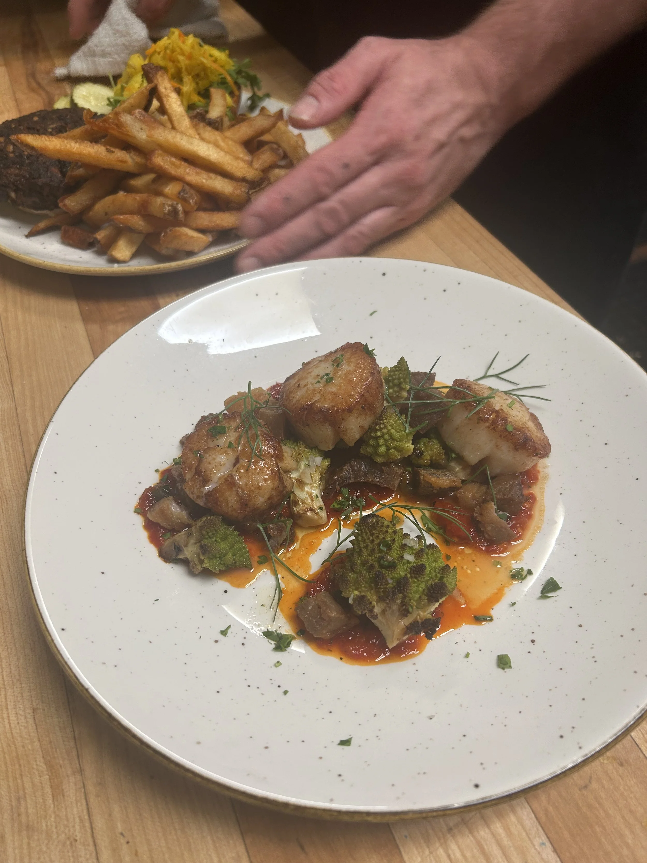 A white plate with seared scallops and roasted Romanesco broccoli on a red sauce garnished with herbs. In the background, there is a plate of French fries and a burger patty with a small side of yellow coleslaw. A person's hand is reaching for the fr