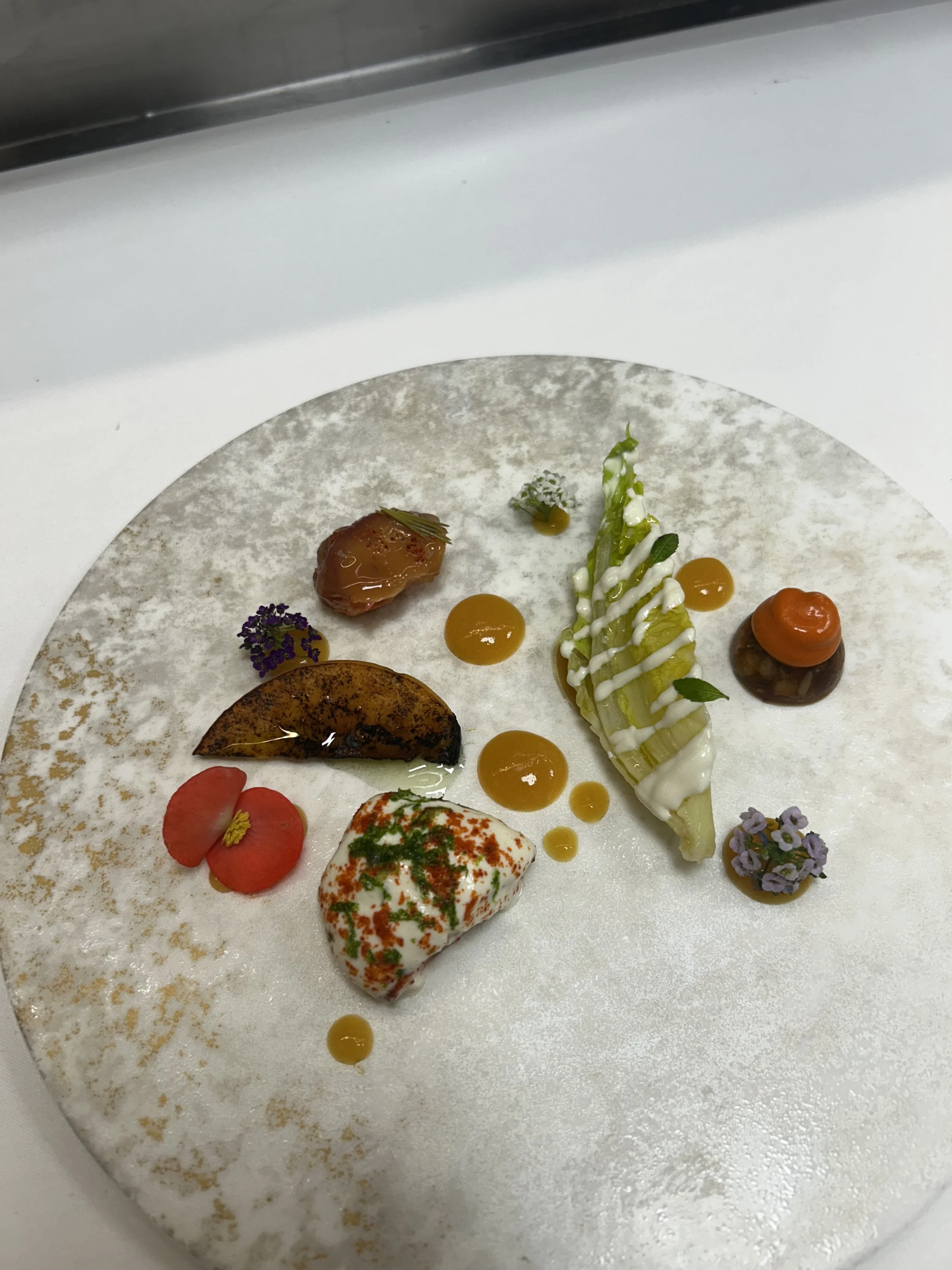 A creative and artistic plated dish featuring various small food items and garnishes on a textured white plate.