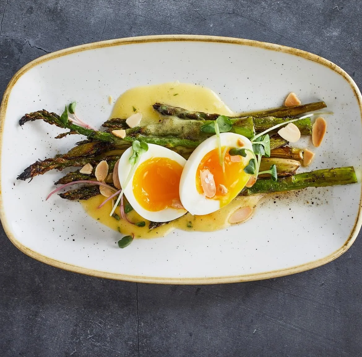 Grilled asparagus topped with a halved soft-boiled egg, microgreens, and sliced almonds on a white oval plate.