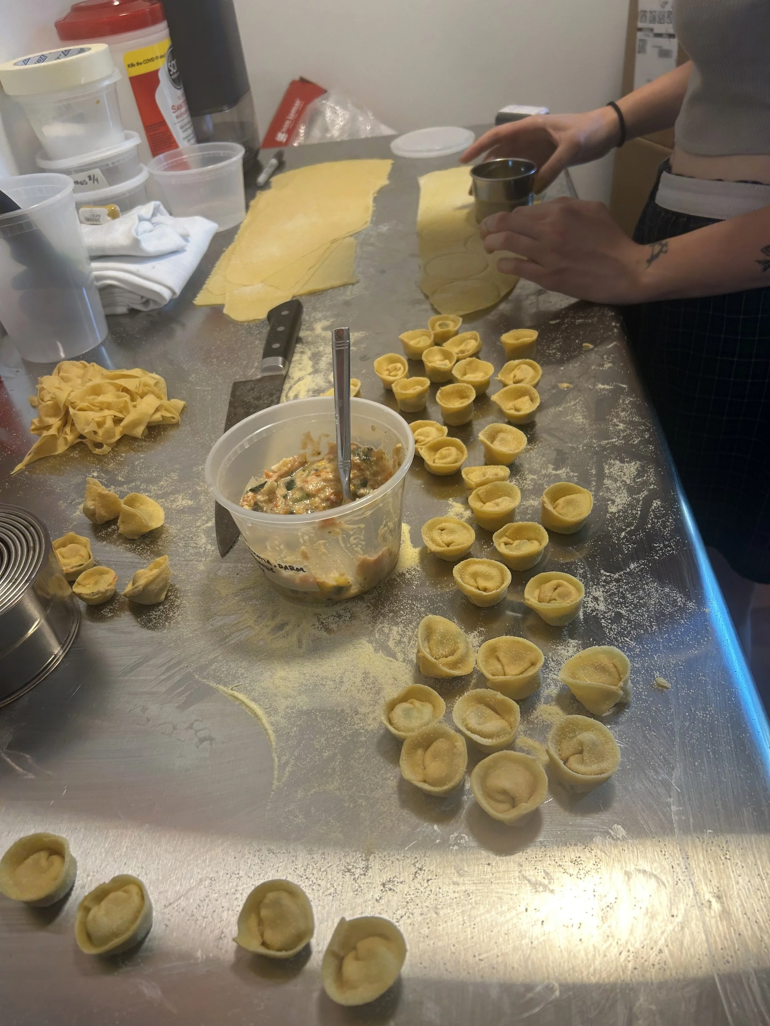 Person making tortellini pasta on a stainless steel table, with dough, filling, and various pasta-making tools.