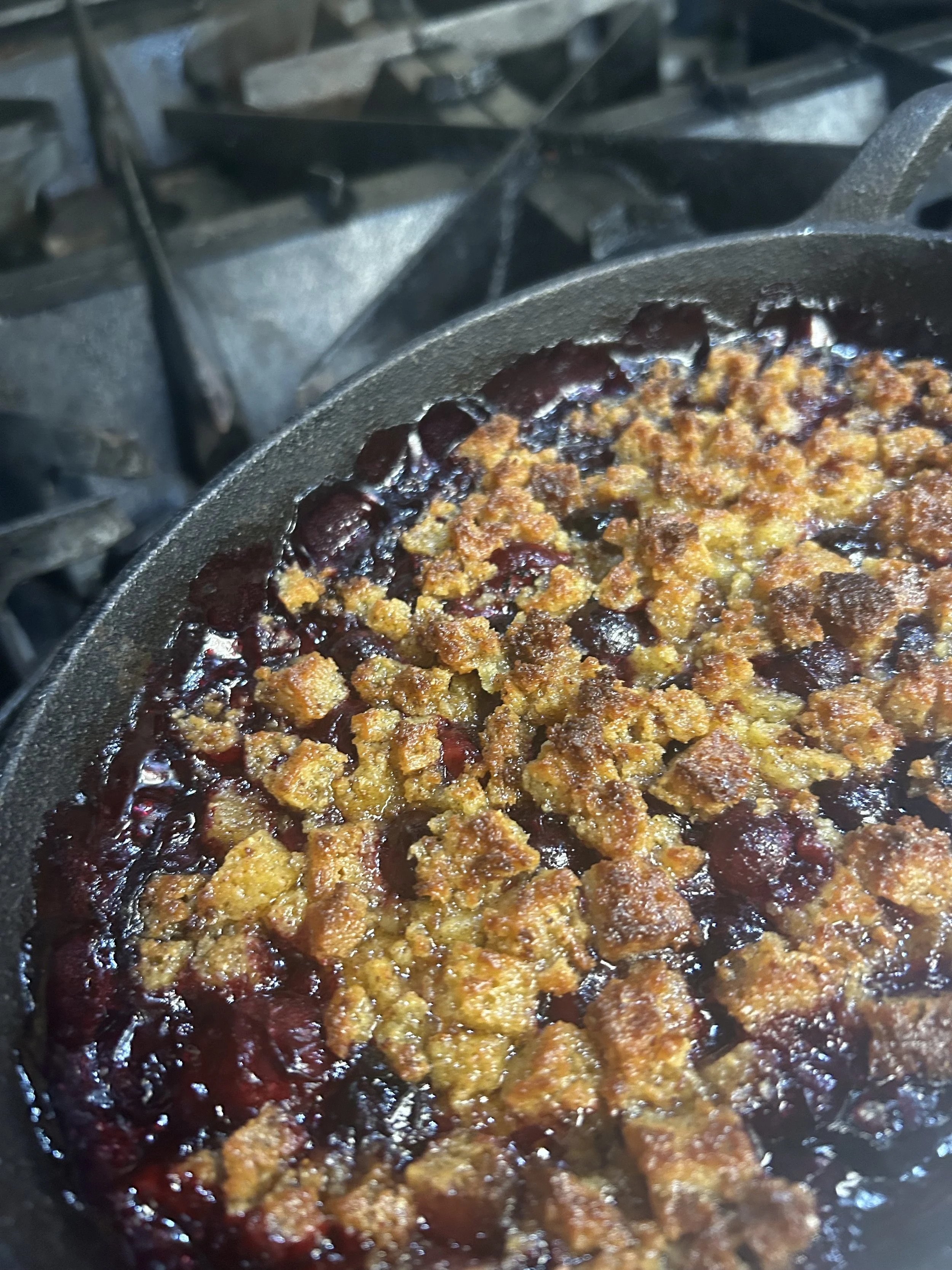 Close-up of a skillet with cooked fruit filling topped with a crumbly golden-brown crust, likely a berry cobbler in a kitchen stove.